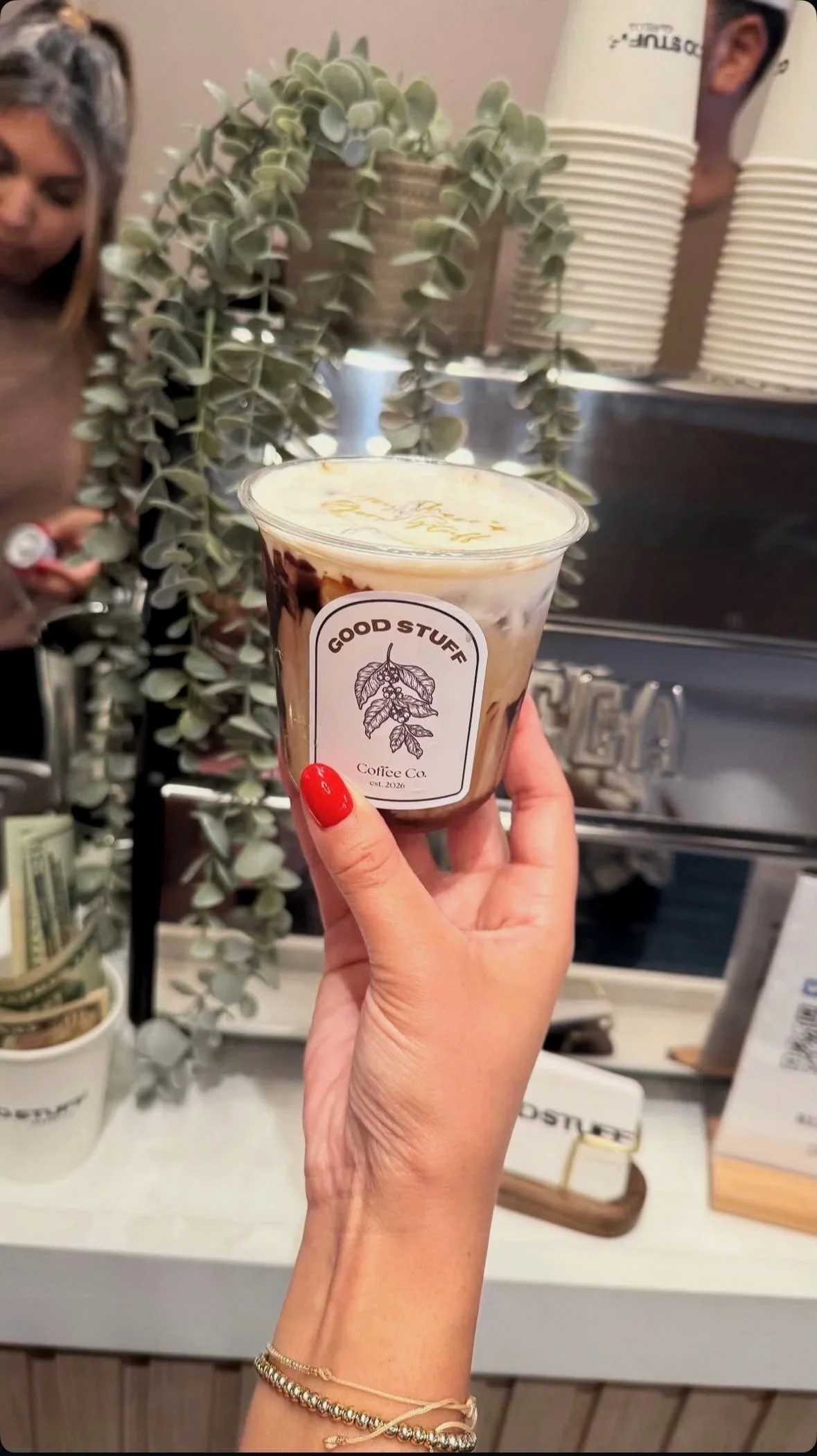 A close-up of a hand with red nail polish holding a plastic cup with a layered beverage, with a label reading 'Good Stuff Coffee Co.' in front of a counter with plants and coffee equipment.