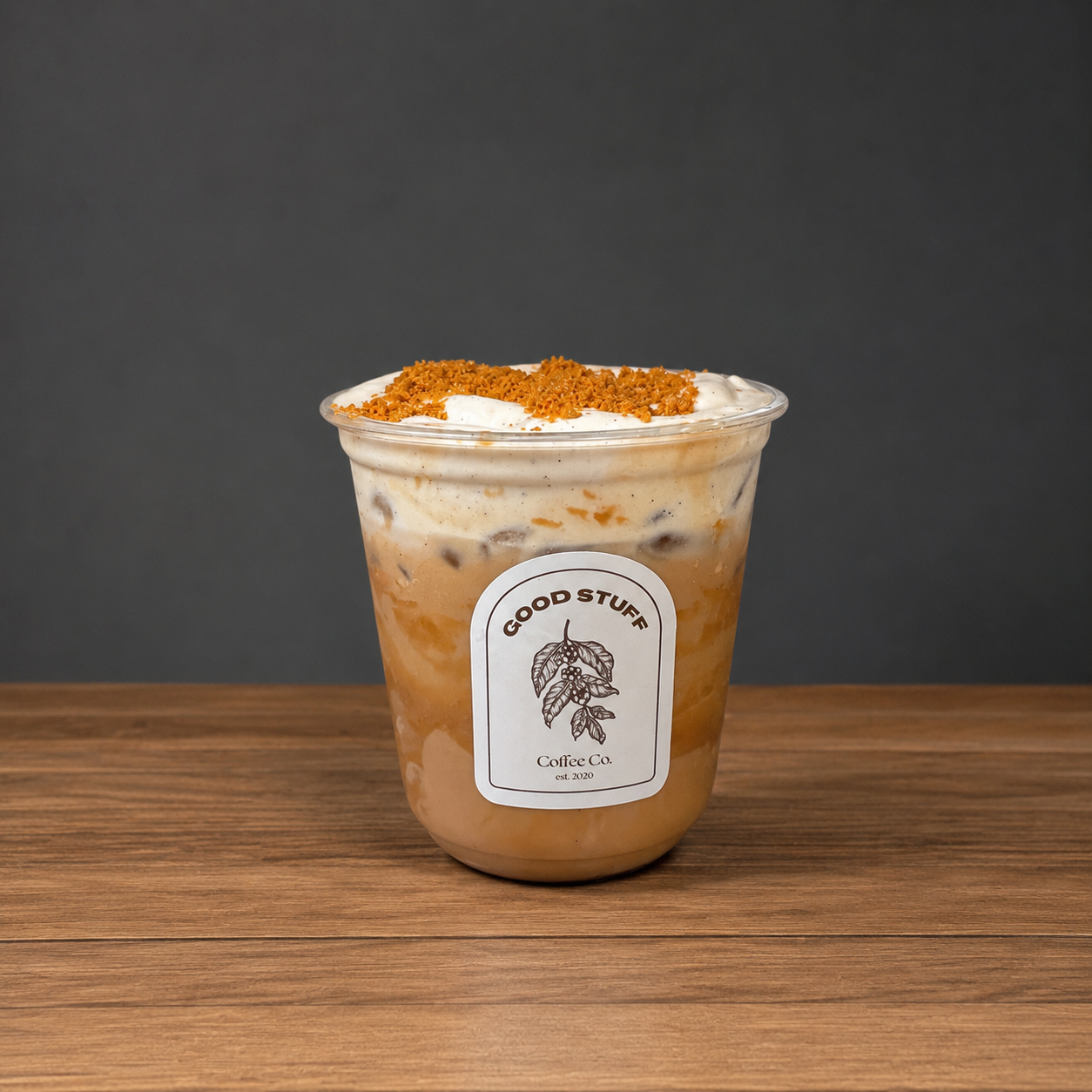 A plastic cup filled with iced coffee topped with whipped cream and caramel crumbles, labeled 'Good Stuff Coffee Co.' on a wooden surface against a dark background.