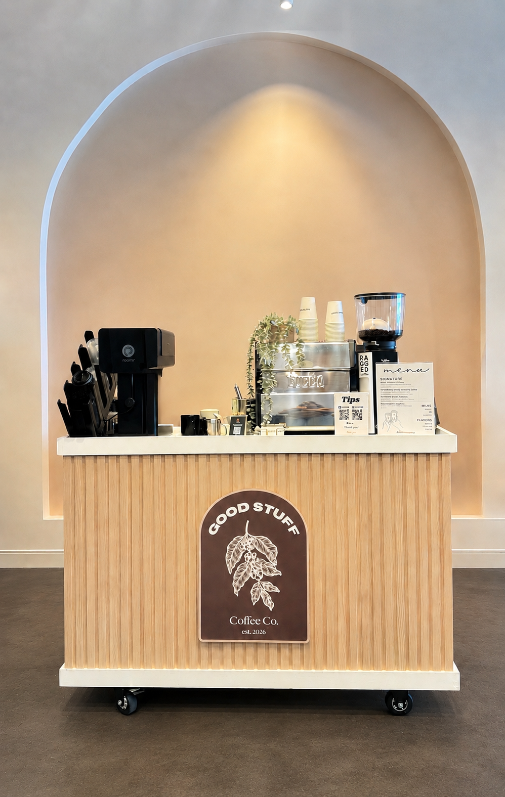 A coffee stand with a light wood panel front, featuring a sign that reads 'Good Stuff Coffee Co' with a coffee leaf illustration, situated in a minimalist space with beige walls and an arch-shaped wall niche.