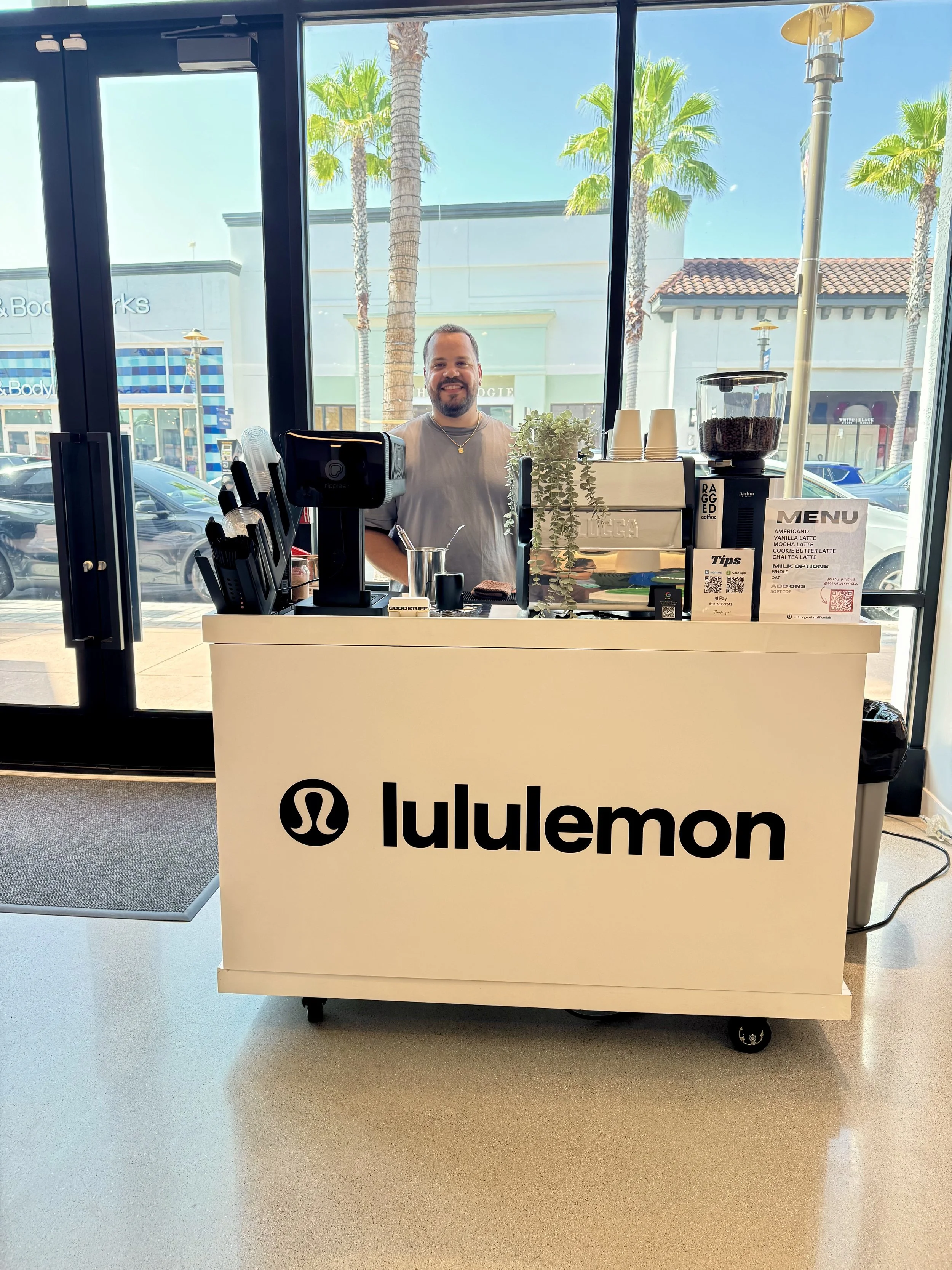 Inside a retail store with large glass windows showing palm trees outside. A man with a beard and gray t-shirt stands behind a white cart with 'lululemon' branding, holding coffee equipment and a small potted plant, smiling at the camera.