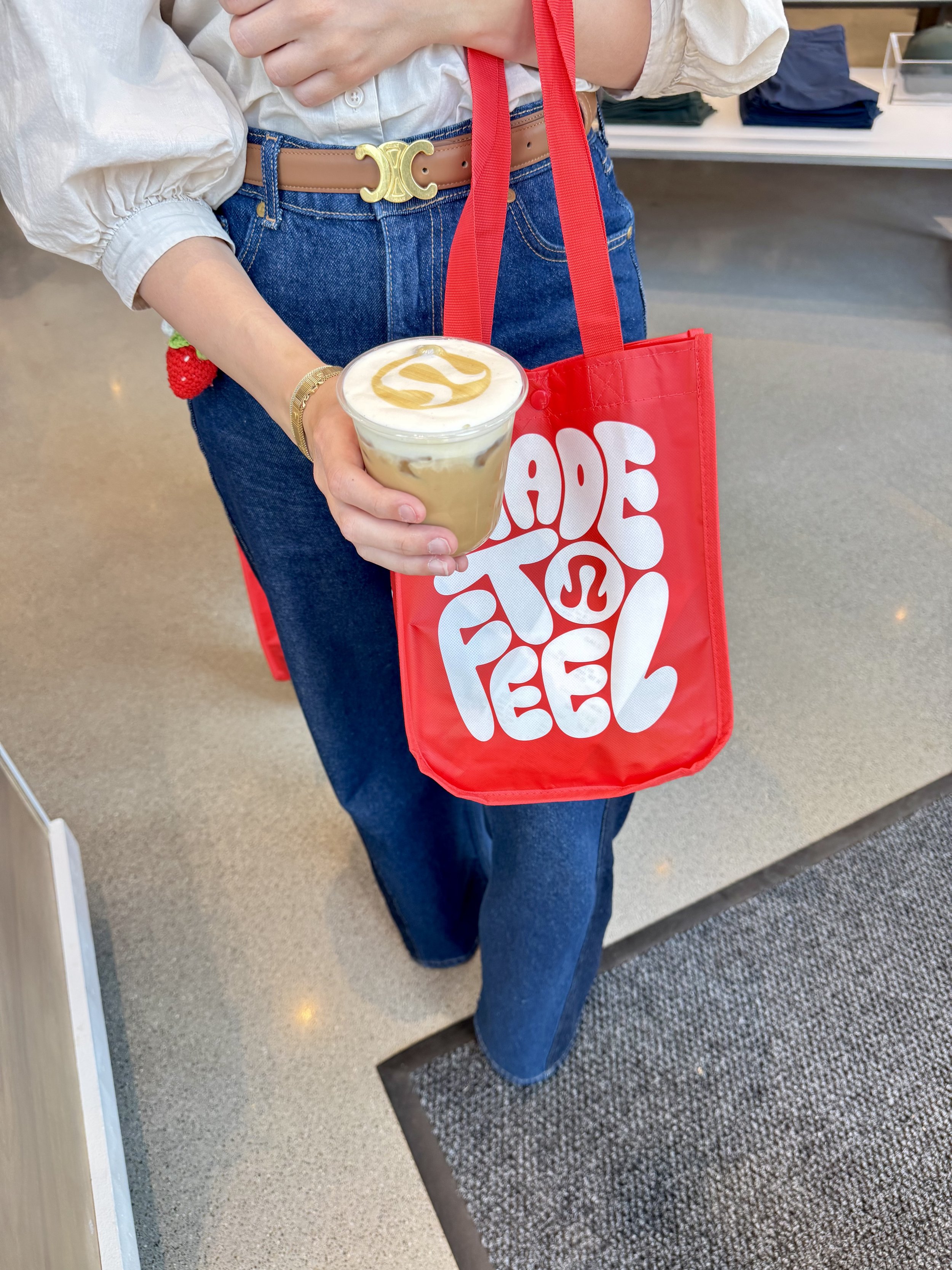 Person holding a cup of coffee with caramel drizzle on top, carrying a red reusable shopping bag with a white logo, wearing a white blouse, blue jeans, a tan belt with a gold buckle, and a gold bracelet, standing on a carpeted and tile floor.