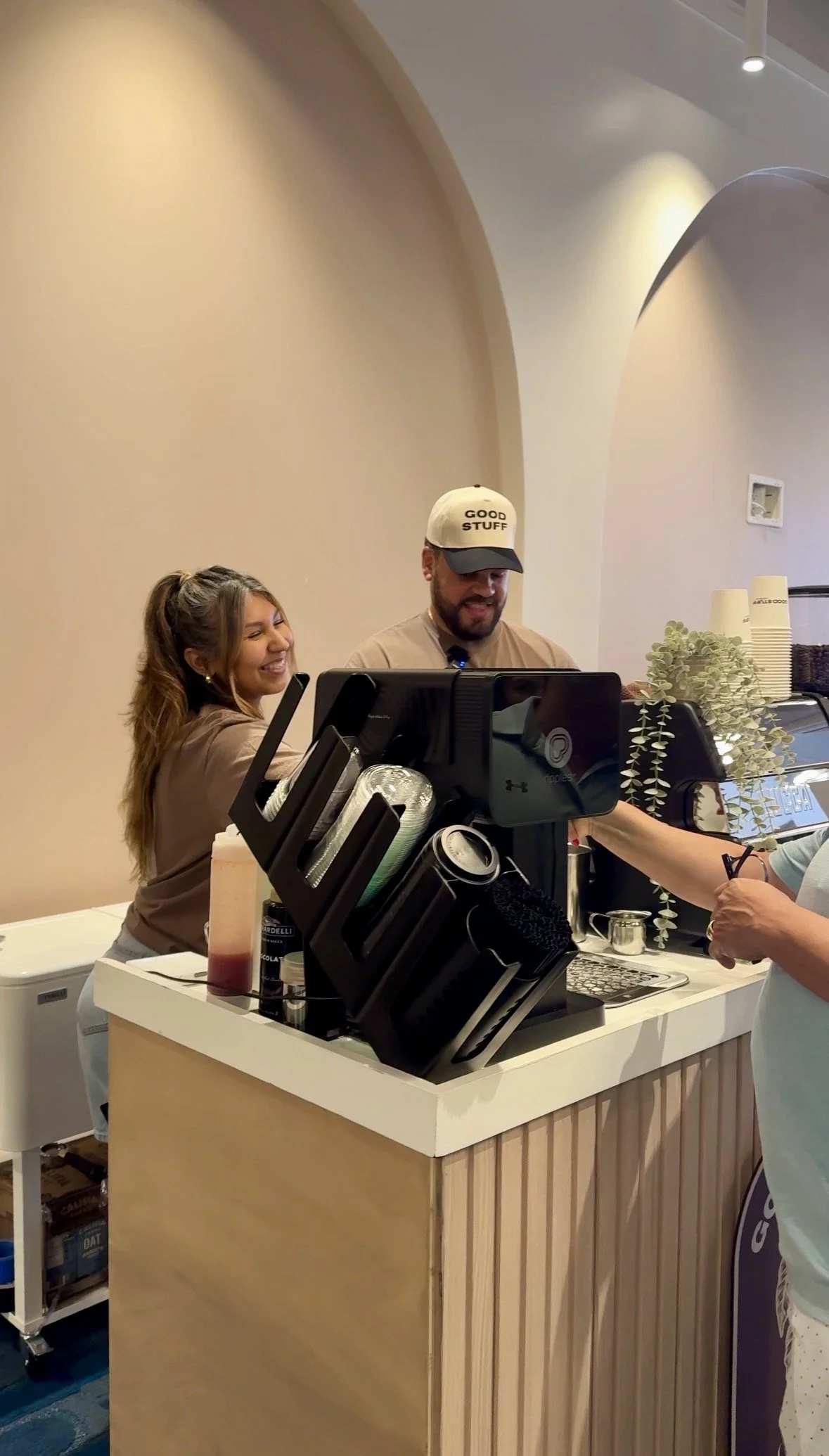Smiling woman and man behind a coffee shop counter. The woman has long brown hair in a ponytail, and the man is wearing a baseball cap that says 'Good Stuff'. There are coffee cups, a cash register, and bottles on the counter.