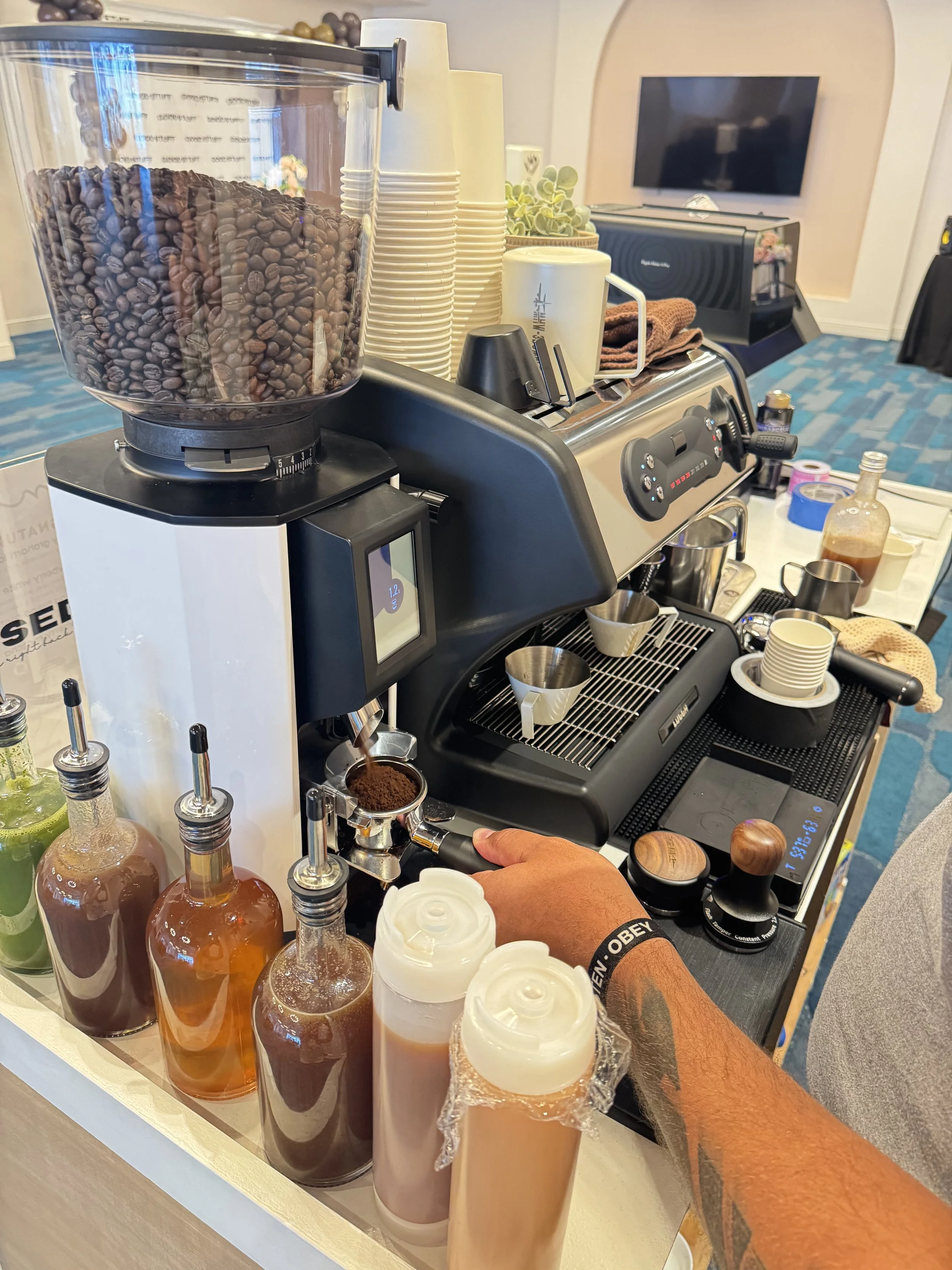 A commercial coffee machine with multiple bottles of flavoring syrup, disposable cups, and a barista's hand operating the machine, set up on a counter in a cafe or event space.