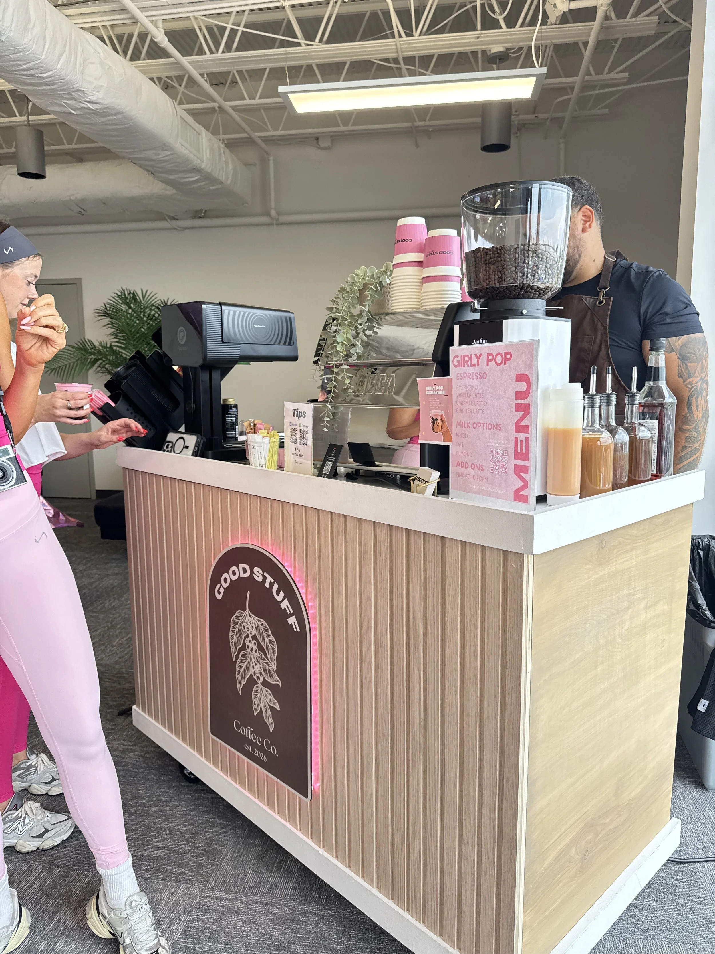 A coffee stand at a store, with a person behind the counter making an espresso, a customer in front holding a pink coffee cup, and a sign displaying coffee options. The stand is decorated with a coffee logo and pink accents.