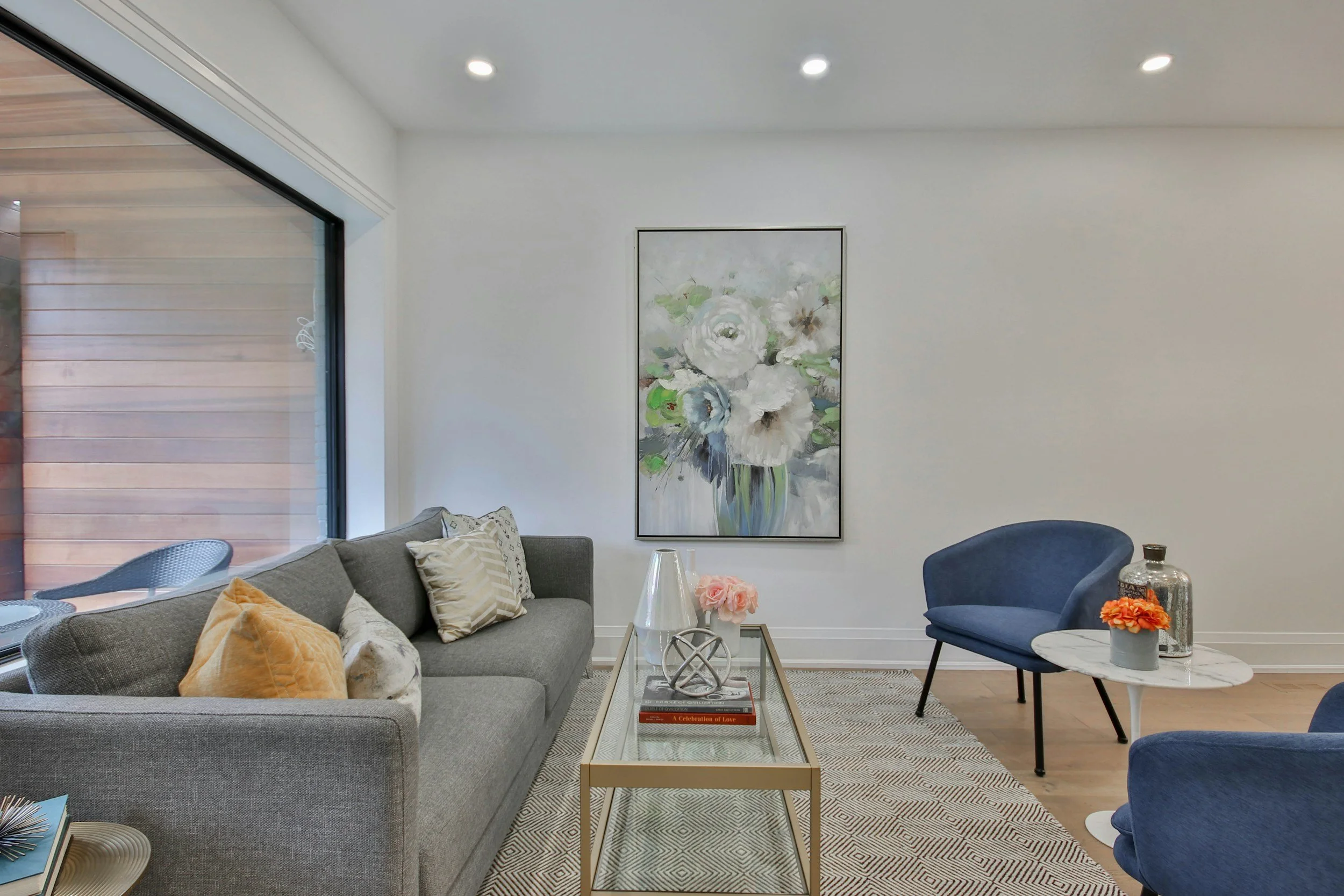 Living room with a gray sofa with beige, white, and patterned pillows, a beige armchair, a blue armchair, a white marble-topped coffee table, a side table with a pink flower arrangement, another small table with a glass jar, a large floral painting on the wall, a window with wooden shutters, and recessed ceiling lights.
