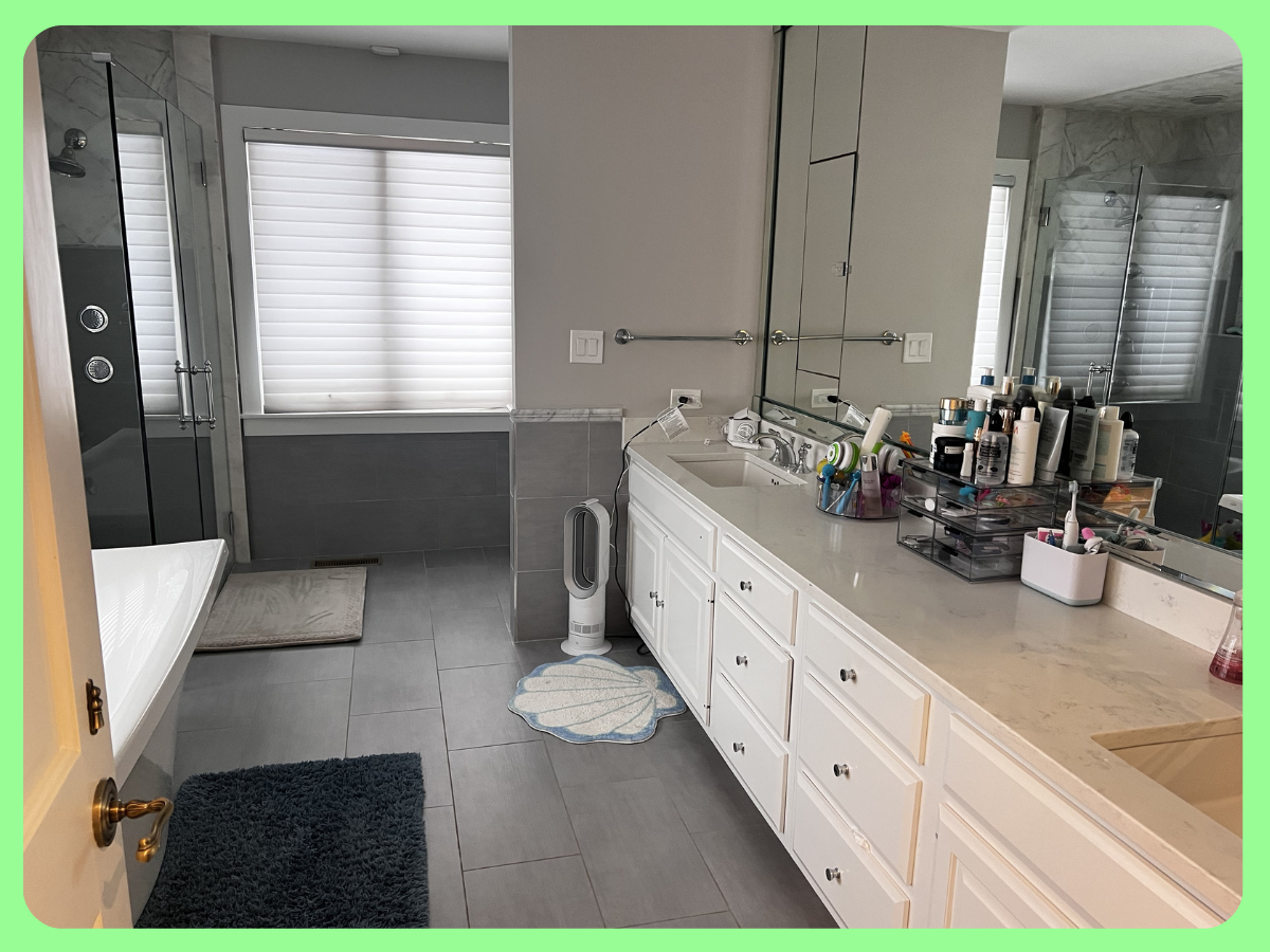 Bathroom with a shower stall, large window with blinds, white vanity with multiple drawers, and various personal care products.