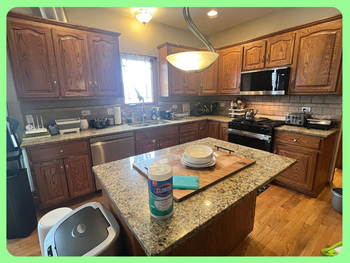 A kitchen with wooden cabinets, granite countertops, a stainless steel dishwasher, and a stove with a microwave above. The kitchen island has stacked bowls, a cutting board, and a container of disinfecting wipes. There are various small appliances an