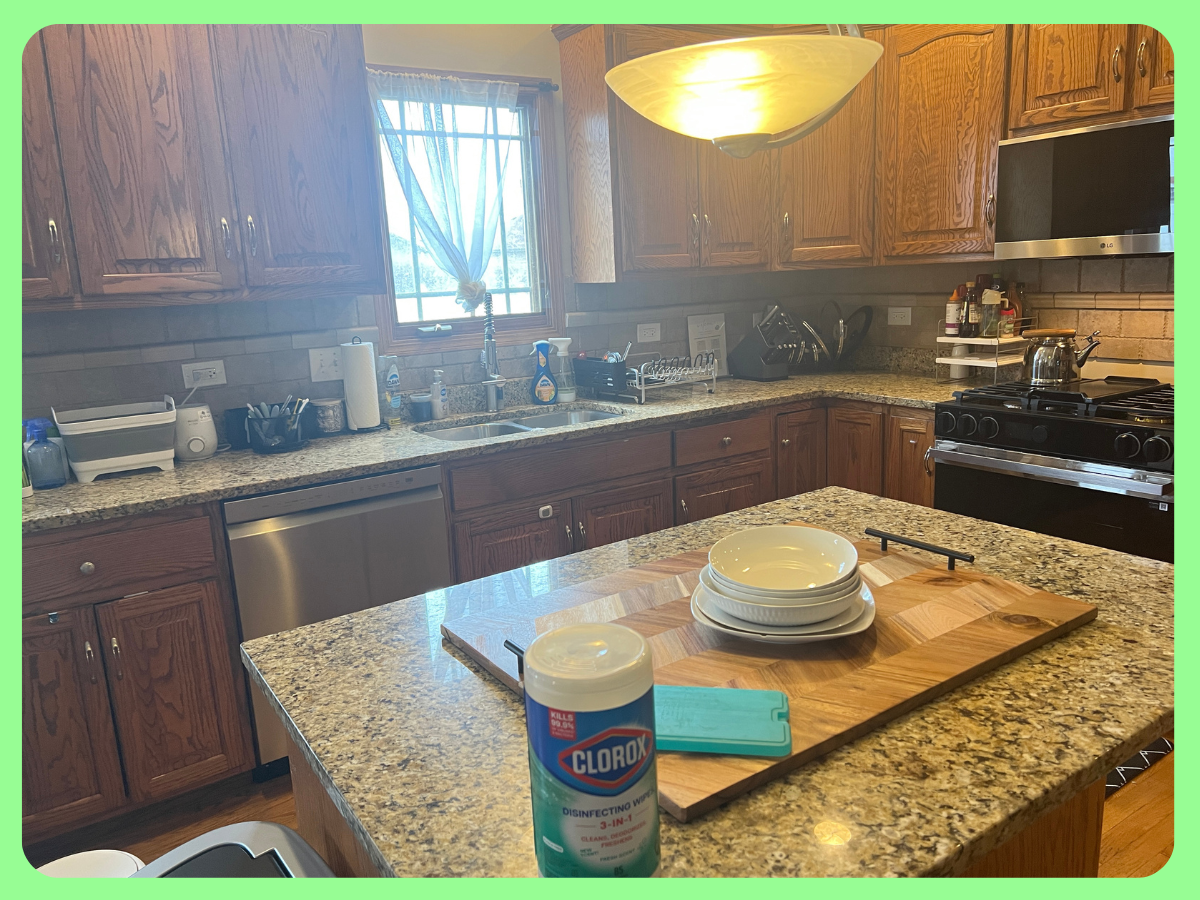 Kitchen with wooden cabinets, granite countertops, a window with curtain, and various appliances and items. Contains a chopping board with bowls, a container of disinfecting wipes, a microwave, a stove with a kettle, and dish soap near the sink.