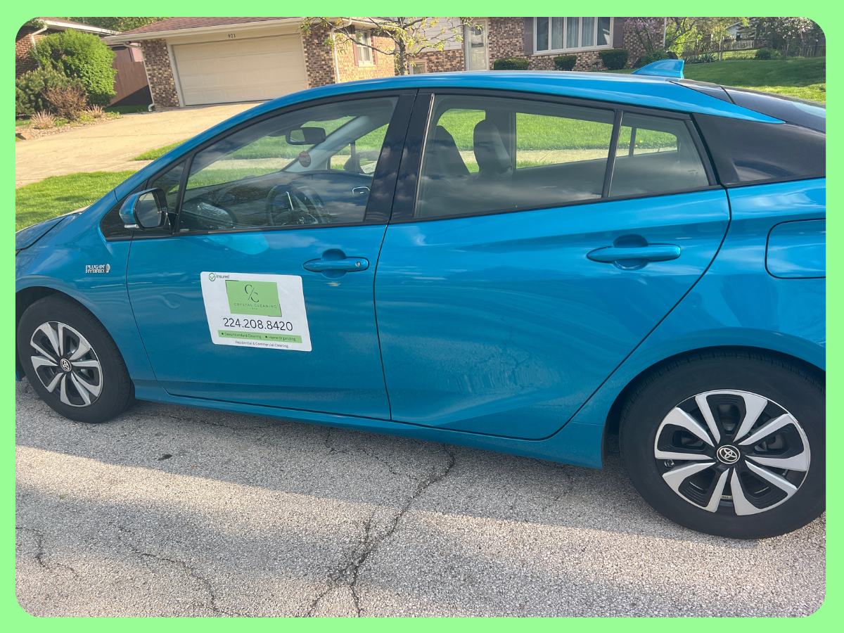 Blue hybrid Toyota Prius parked on a residential street with a house and grass in the background. A door hanger sign with contact information is attached to the driver side door.