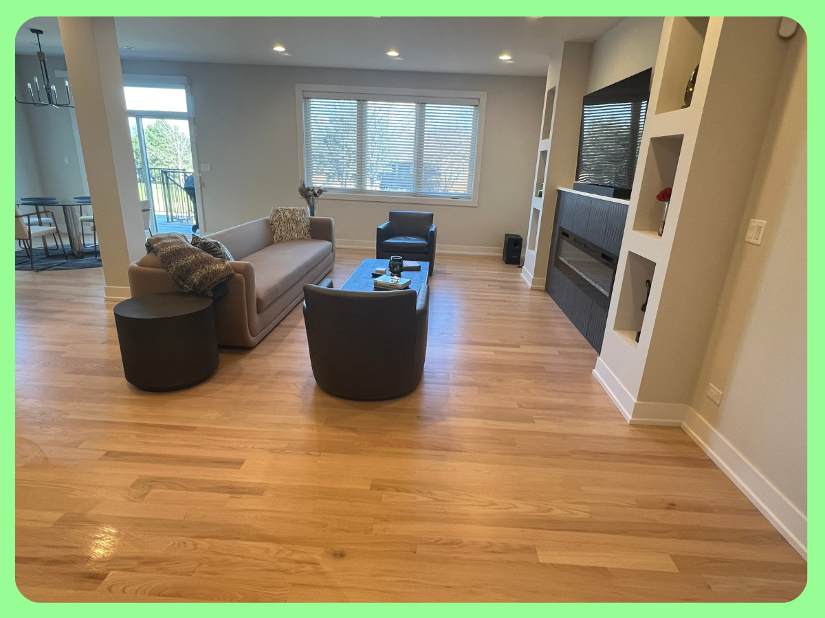 Living room with a beige sofa, black armchair, black ottoman, and a small black side table. There is a large window with blinds, a fireplace with a dark grey surround, built-in shelves, and a mounted TV above the fireplace. Hardwood flooring is visib