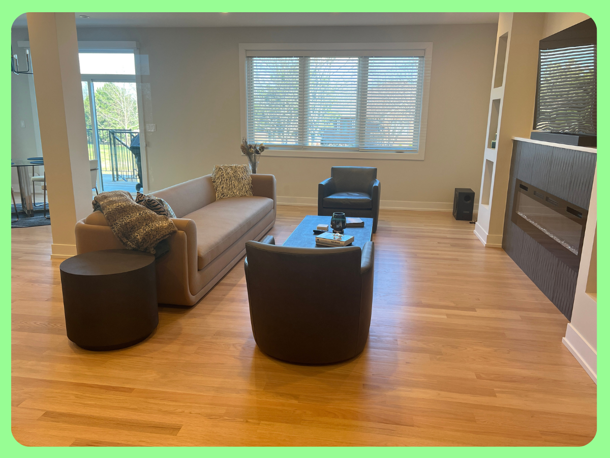 Living room with sofa, two armchairs, coffee table, fireplace, and large window with blinds, with a balcony visible through sliding doors.