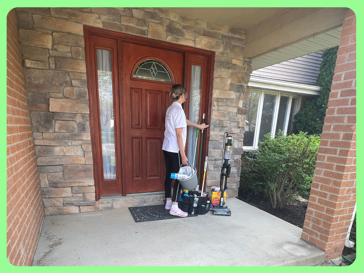 A woman standing on a porch in front of a red front door, organizing cleaning supplies and tools.