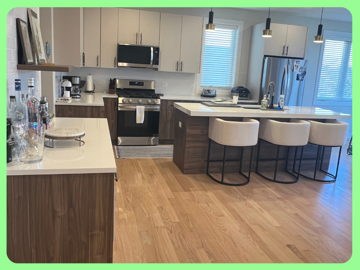 Modern kitchen with white and wood cabinetry, stainless steel appliances, and a kitchen island with four beige bar stools. Hardwood floors and three pendant lights above the island.
