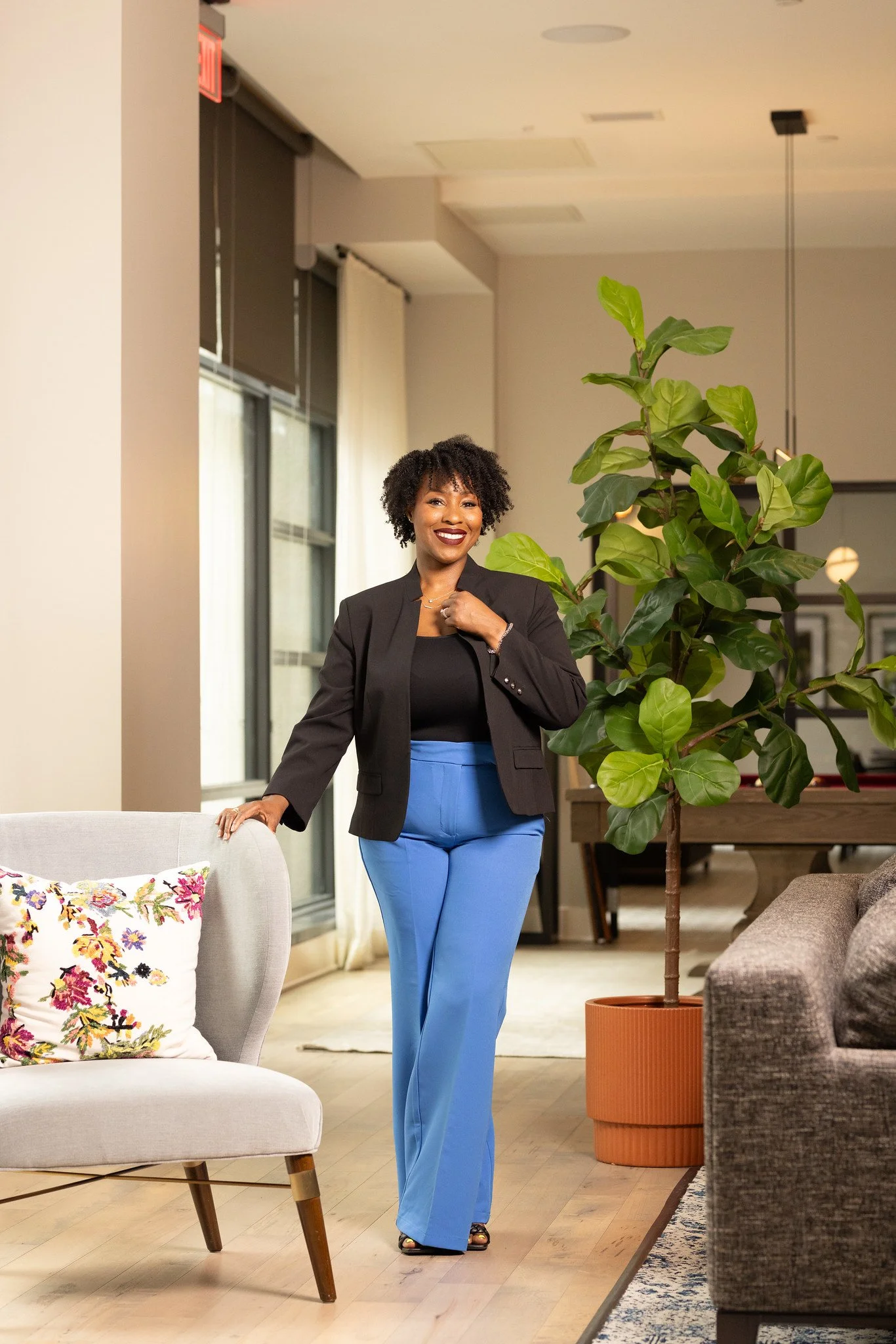 A woman standing in a modern living room, smiling, wearing a black blazer and blue pants, with a large green plant nearby.