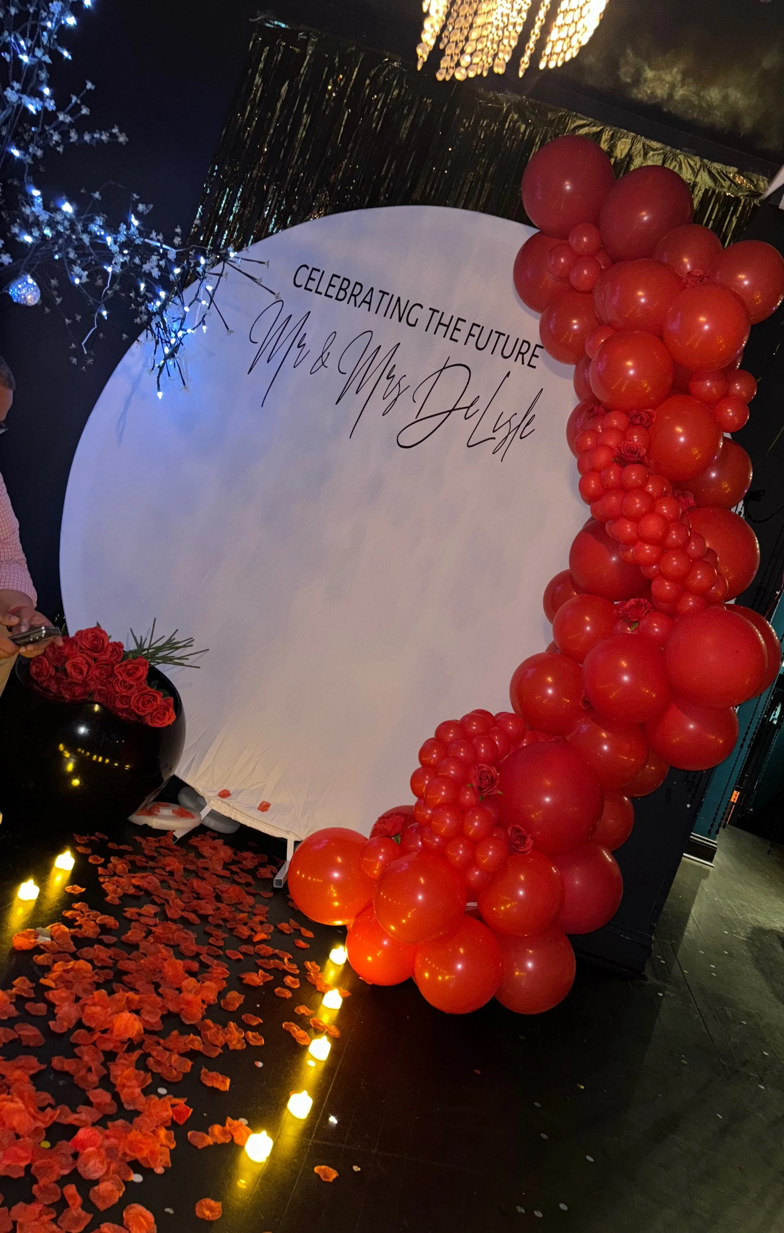 Organic Balloon Arch with red balloons arranged along one side of a circular backdrop, decorated with rose flowers in a black vase, illuminated with string lights, and covered with red flower petals on the floor. 