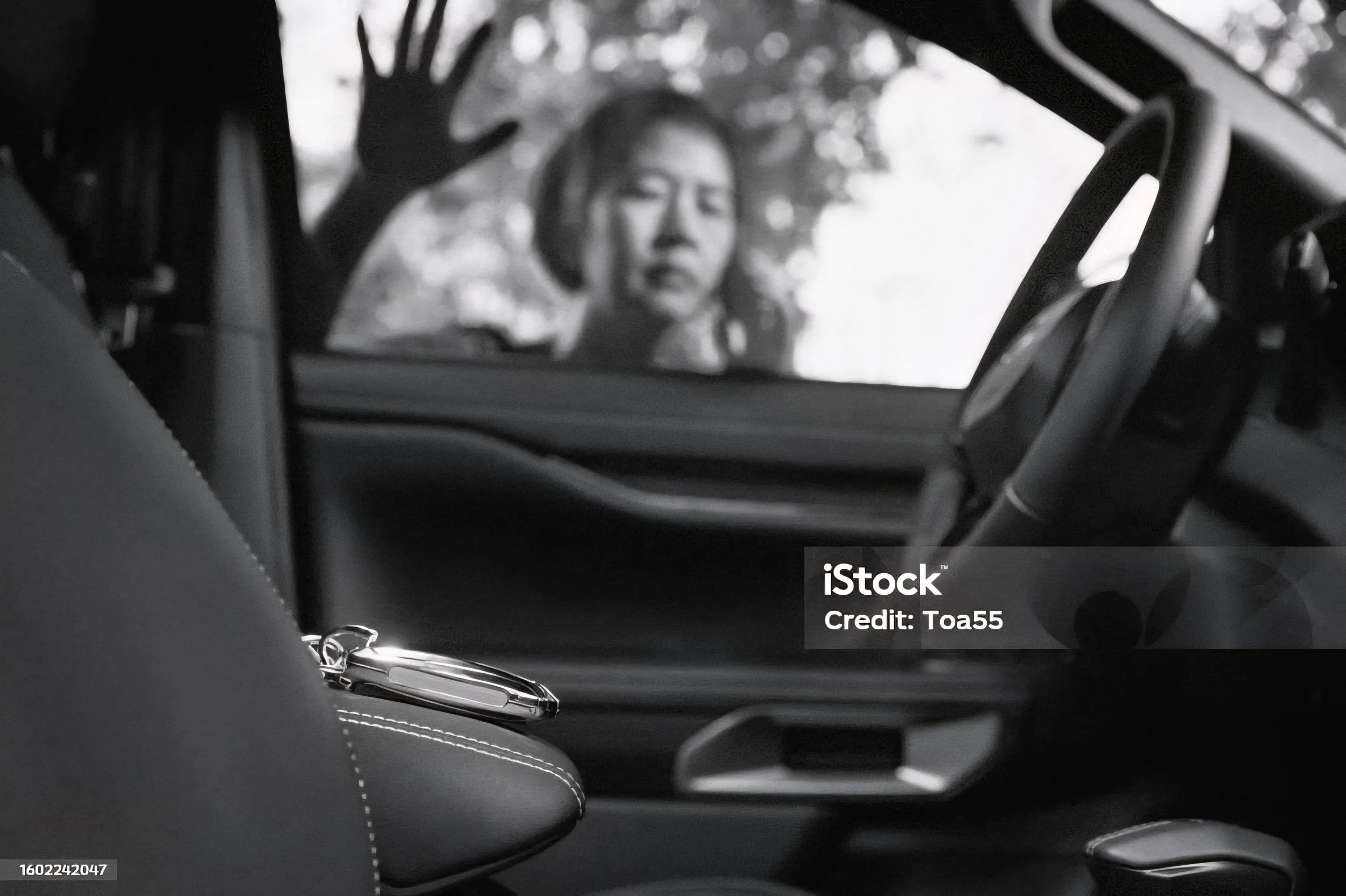 A woman standing outside of a vehicle, looking through the windshield at the camera. The photo is in black and white, with the woman’s facial expression appearing serious and the background featuring trees.
