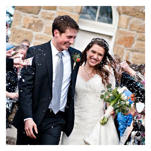 A bride and groom holding hands, smiling at each other, in a field at Chatauqua in Boulder. The bride wears a white strapless wedding dress, holding a bouquet of white flowers. The groom wears a light gray suit with a white shirt and boutonniere.