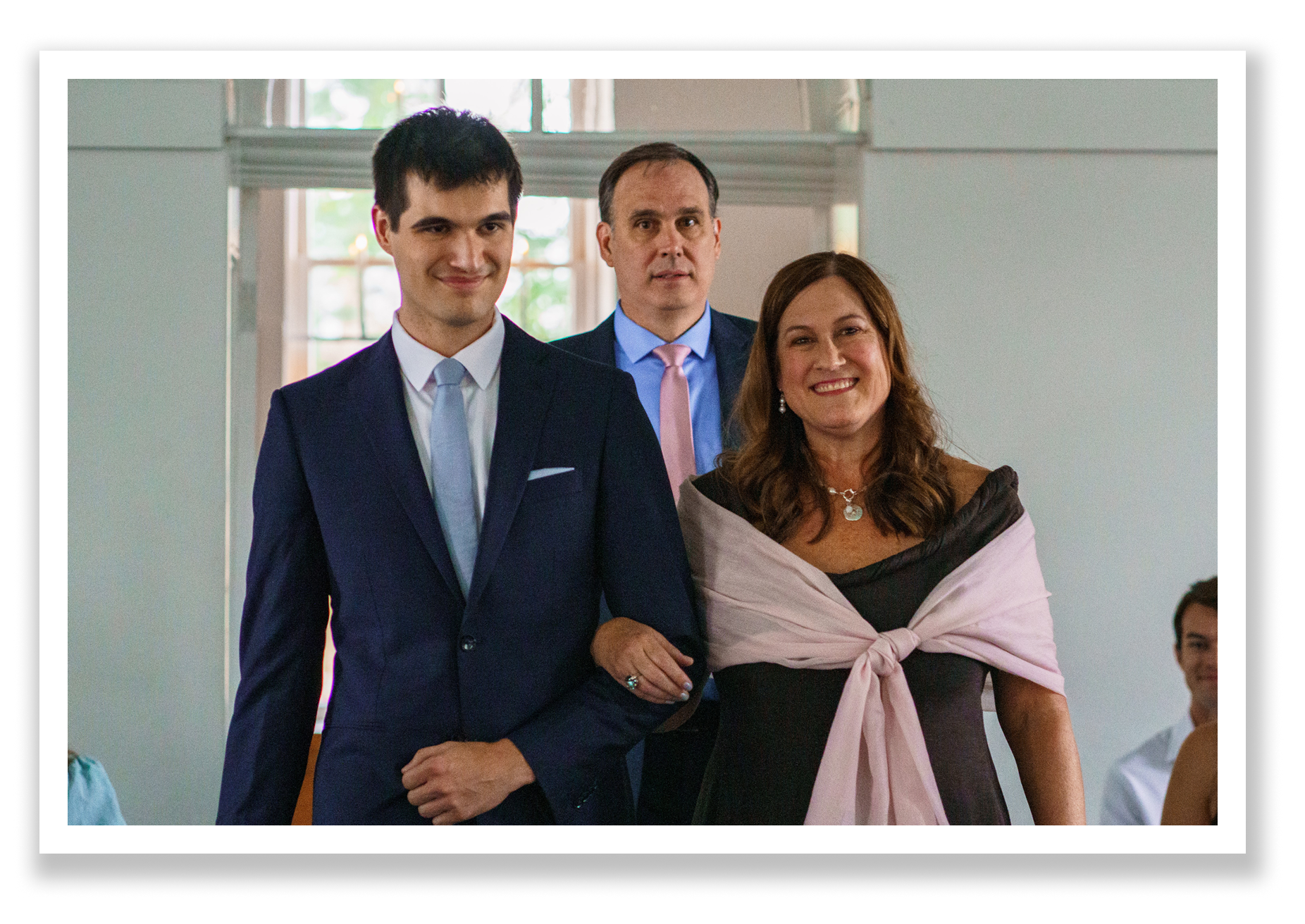 Three people, two men and one woman, who are the groom, his mom, and his dad, are walking down the aisle for a wedding procession. They are dressed formally in a well-lit church sanctuary, smiling at the camera.