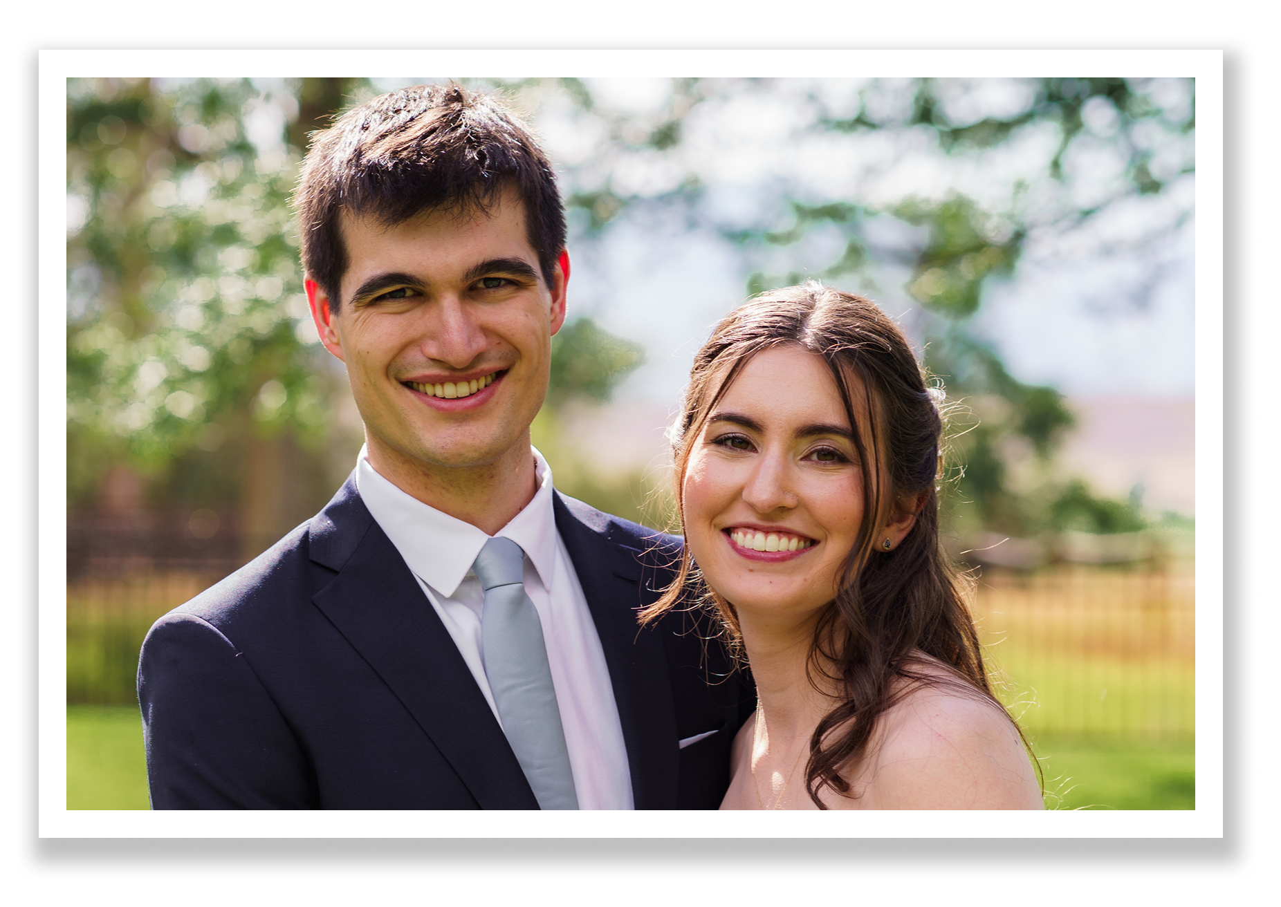 A happy couple who are newlyweds, a man in a suit and a woman with long dark hair in a wedding dress, smiling outdoors with trees and a grassy area in the background.