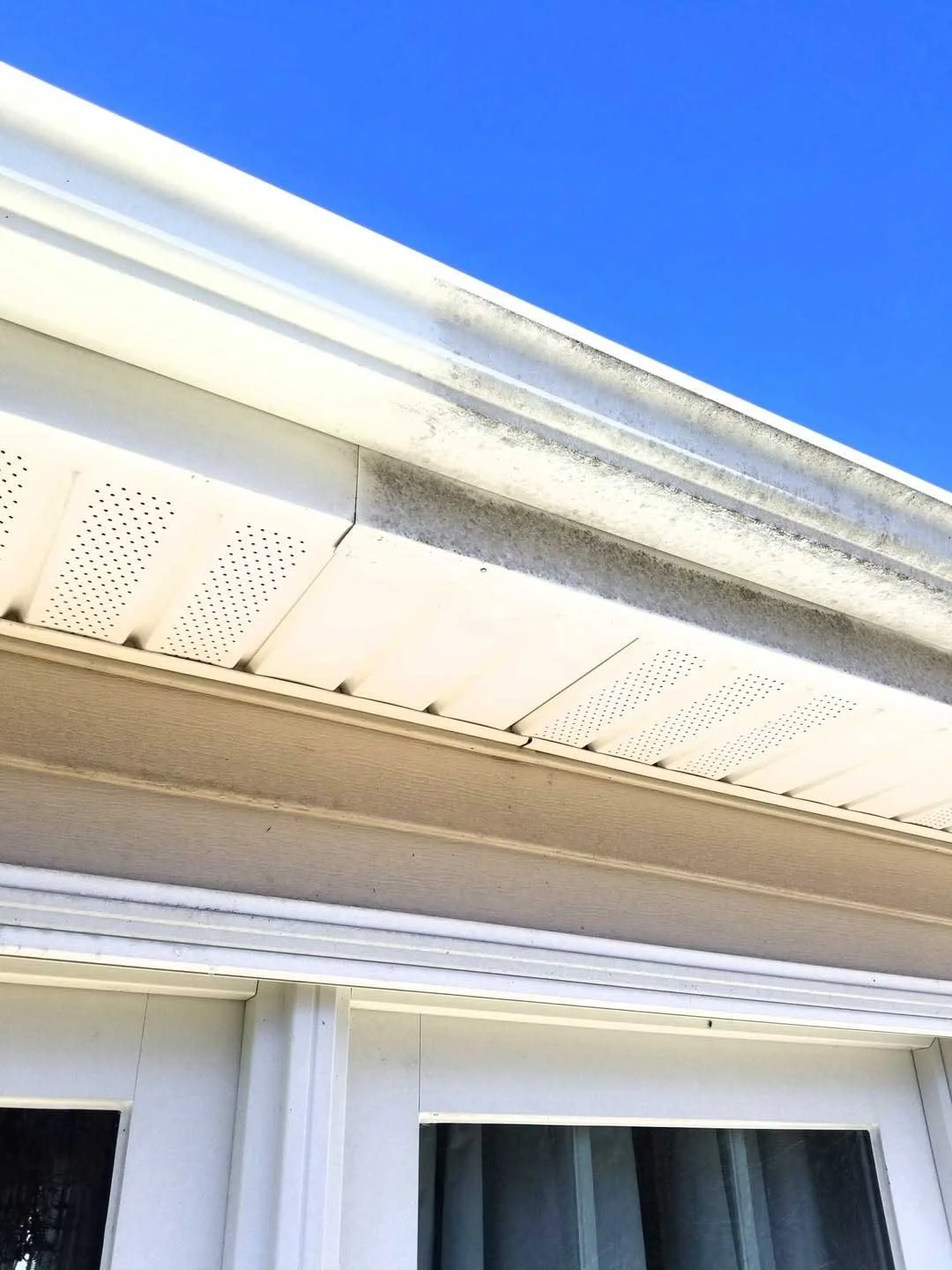 Close-up of a house soffit with white vented panels, showing a before-and-after of glean gutters and some dirt and mold, against a background of a clear blue sky.