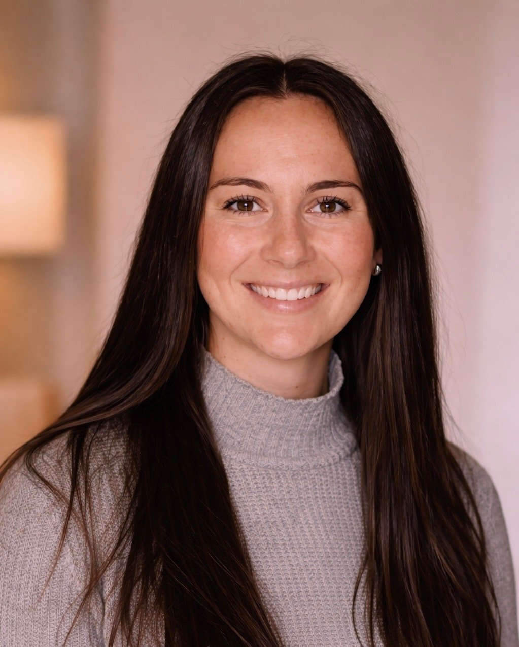A woman with long brown hair, wearing a gray turtleneck sweater, smiling at the camera in an indoor setting.