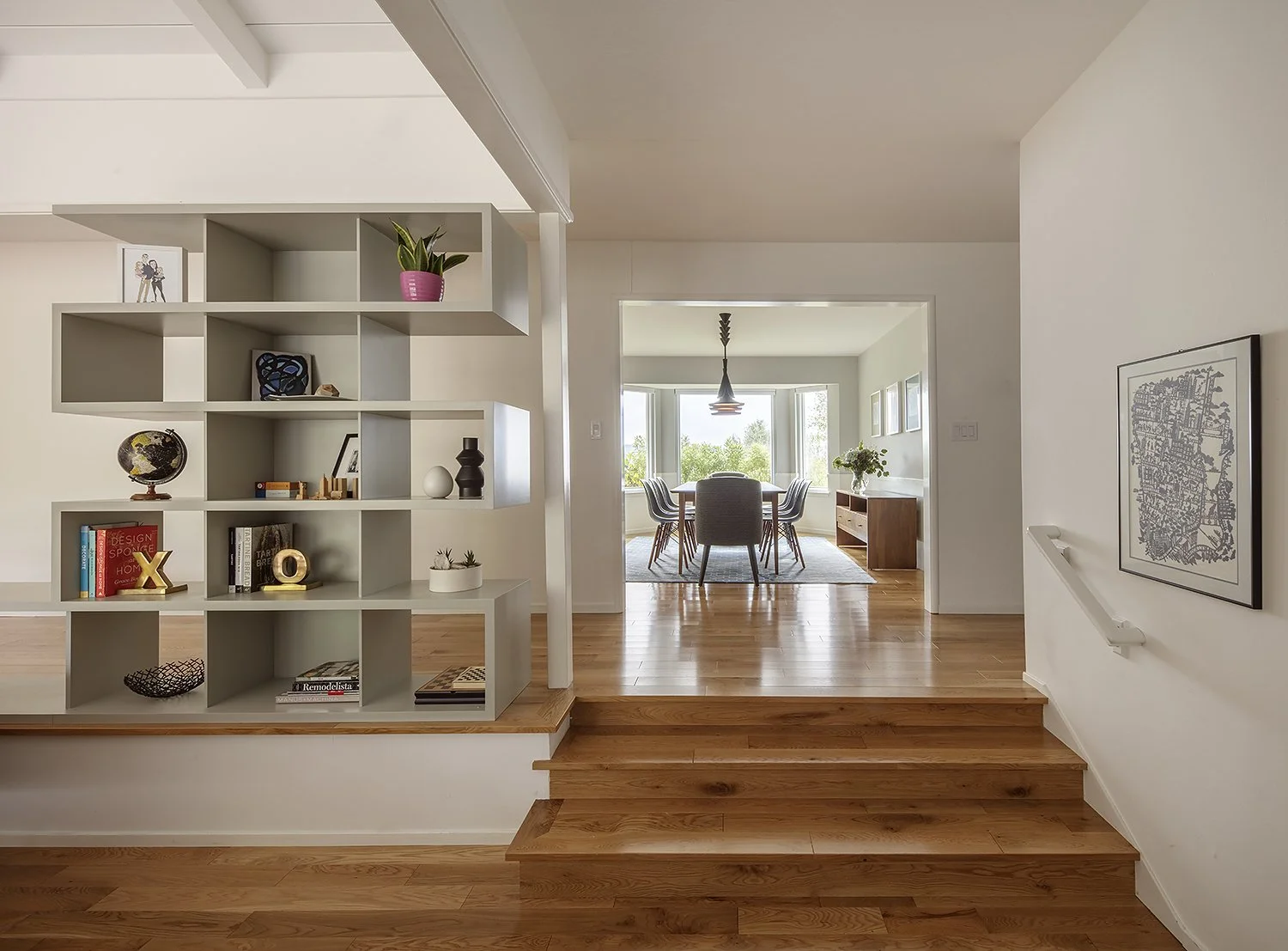 Interior view of a modern home with a white bookshelf, wooden flooring, and a dining area with a table and chairs in the background, large windows with natural light.