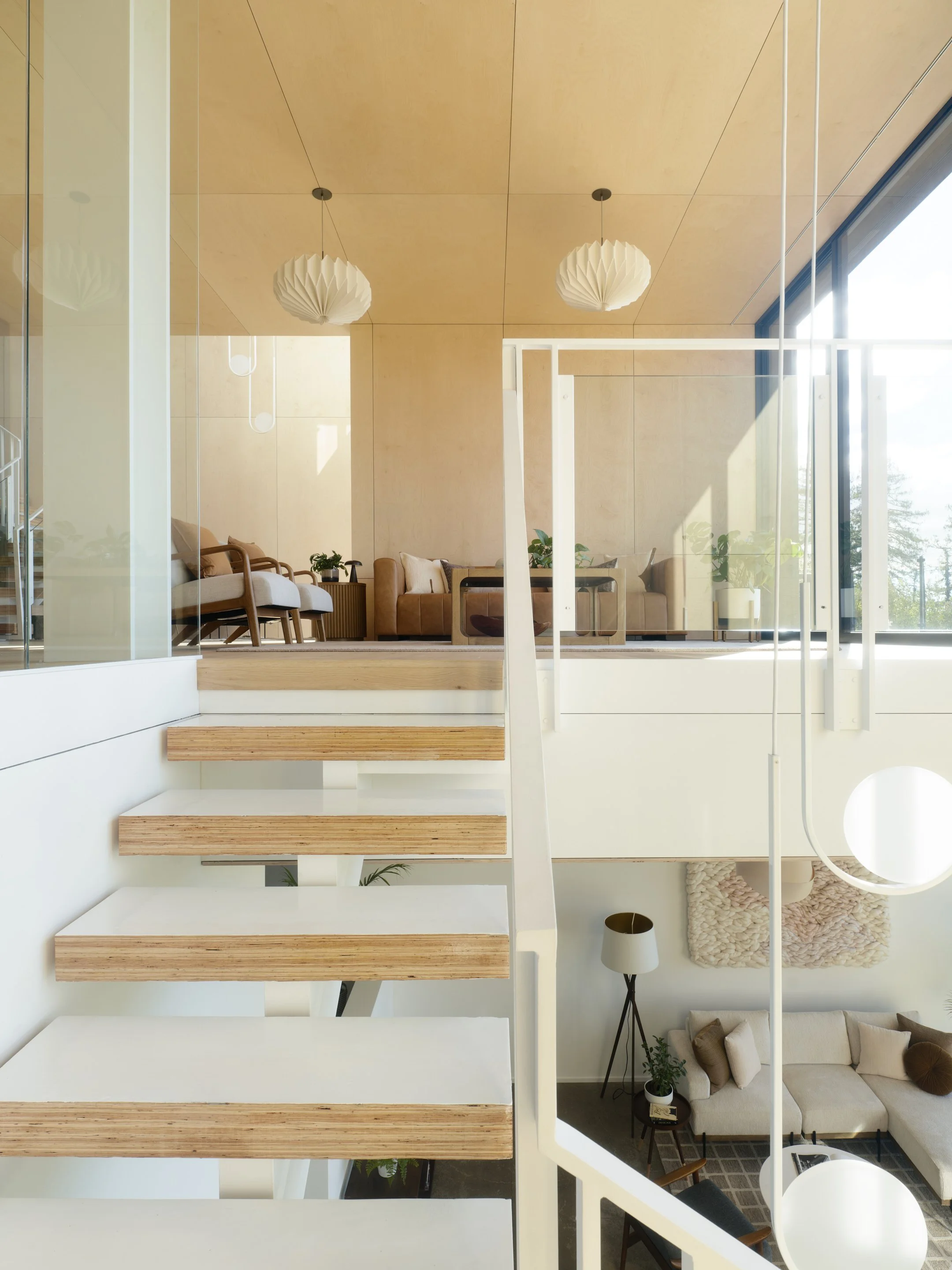 Modern, open-concept living space with wooden stairs, a sitting area with white and brown sofas, plants, a tall lamp, and large windows letting in natural light, viewed from above.