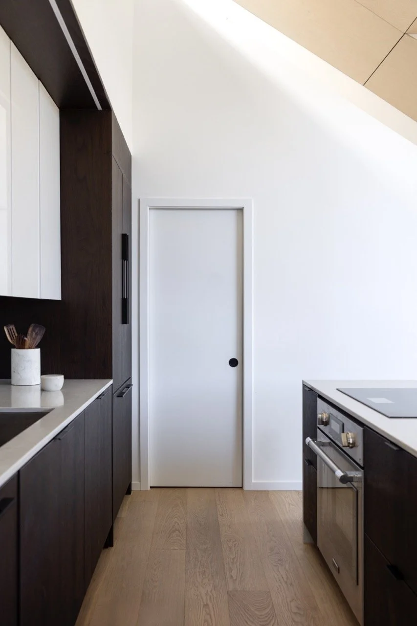 Modern kitchen with black lower cabinets, white upper cabinets, a white countertop, and a white door with a black handle on a sloped ceiling, wood flooring, and white walls.