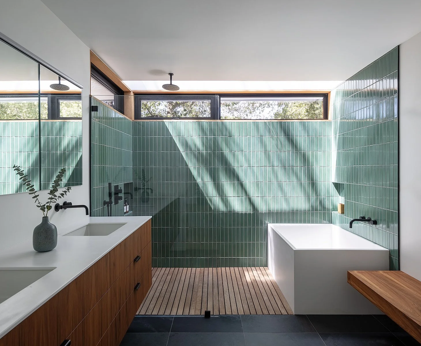 Modern bathroom with green tiled wall, white sink, wooden cabinets, black fixtures, and a bathtub with a wooden side table, natural light from high windows, and a potted plant.