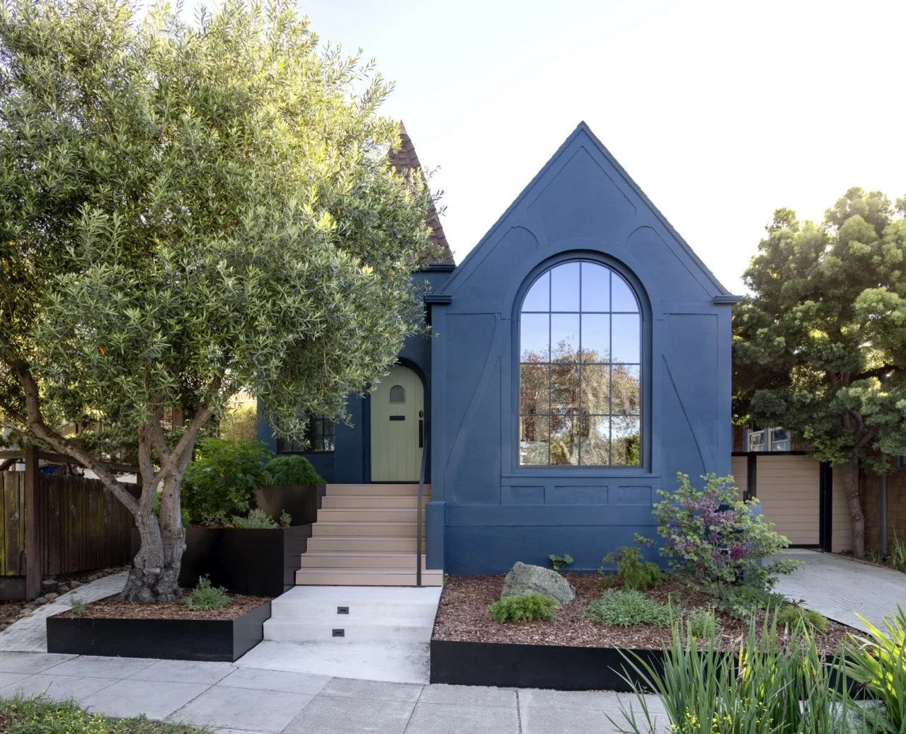 A modern blue house with a large arched window, front stairs, and a front yard garden with trees and plants.
