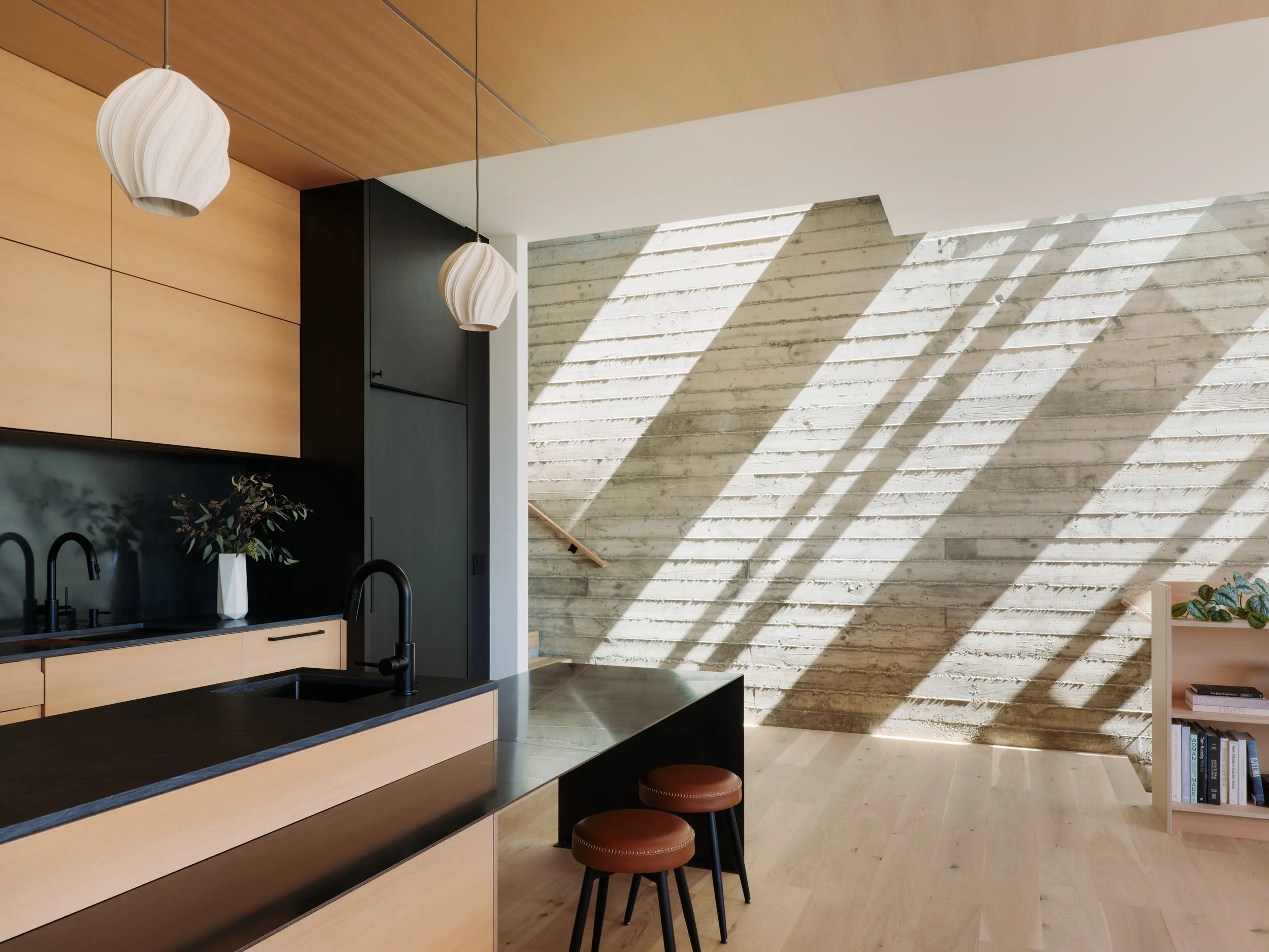 A modern kitchen with wooden cabinets, a black countertop, and two pendant lights hanging from the ceiling. There are two round stools with brown seats in front of the counter. The wall features a textured, light-colored stone design with sunlight ca