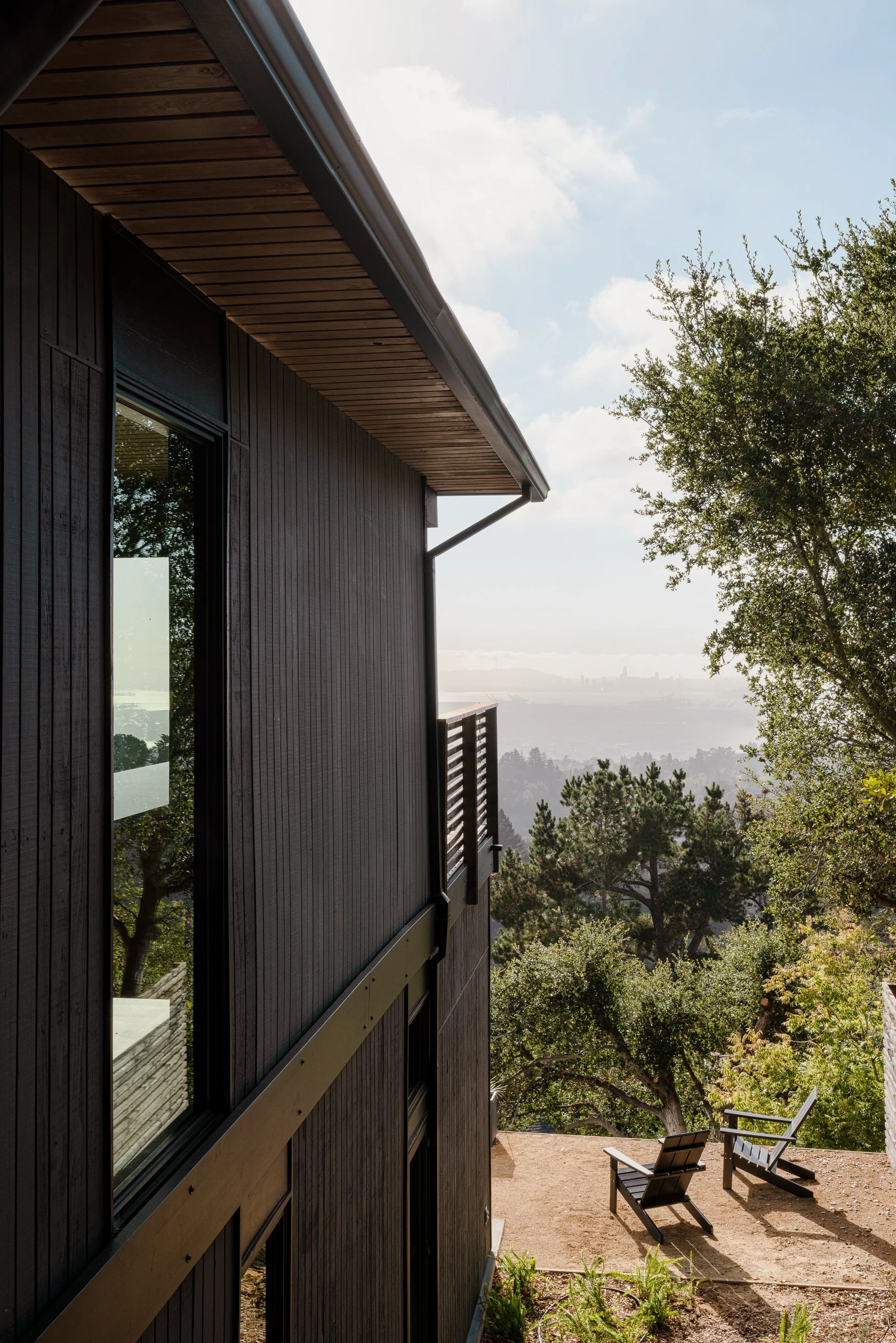 A modern house with dark wood exterior and large windows, overlooking a scenic view of trees and hills in the distance, with two outdoor chairs on a dirt patio.