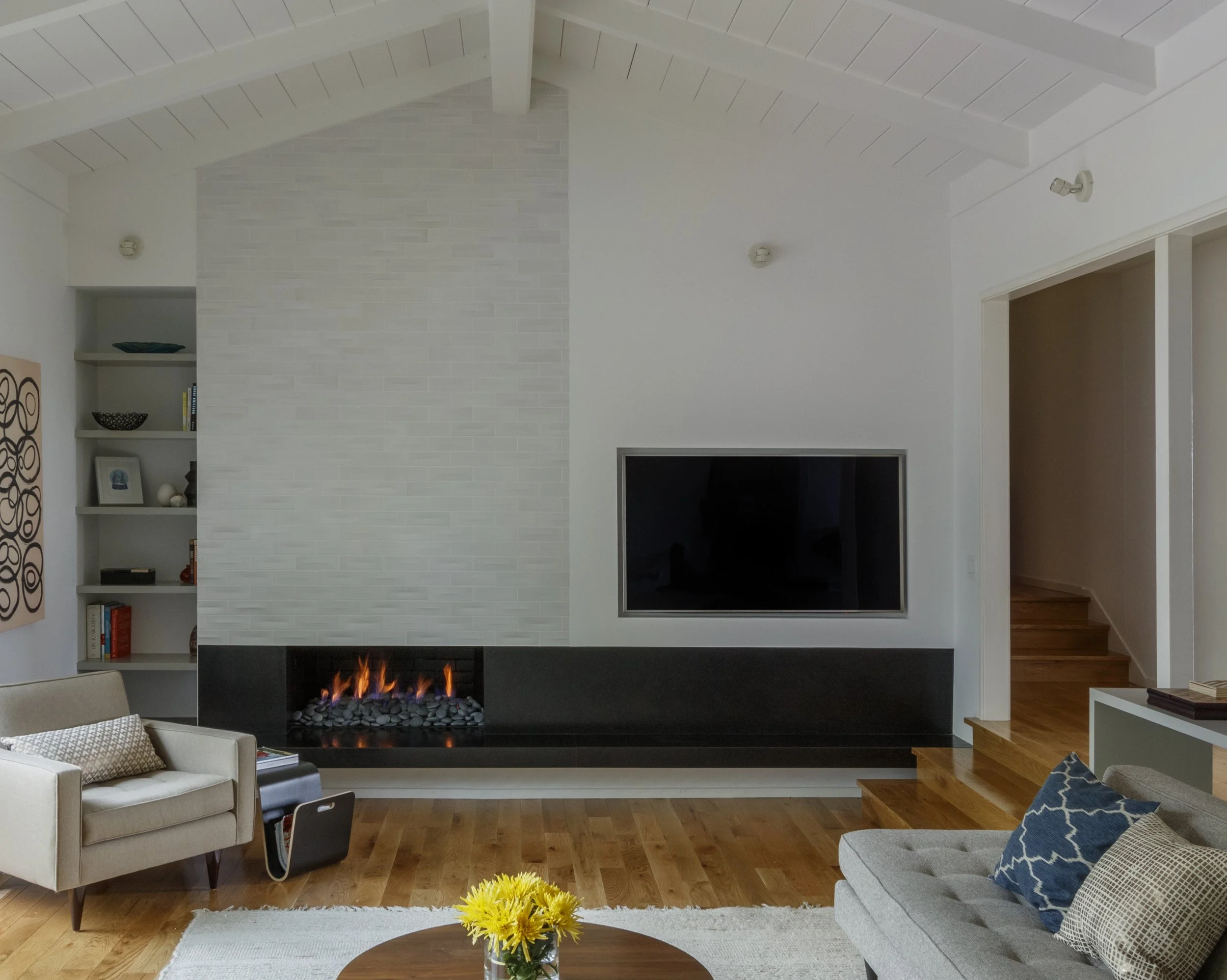 Living room with a modern fireplace, bookshelf, beige sofa, armchair, wooden floors, and a flat-screen TV mounted on the wall.