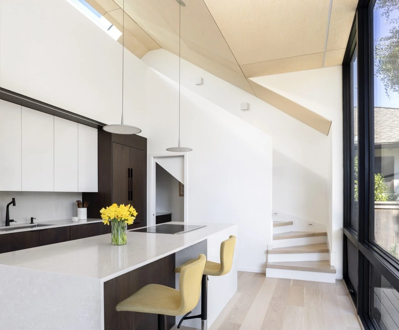 Modern kitchen with white and dark wood cabinetry, yellow chairs, a white kitchen island with a vase of yellow flowers, pendant lights, large windows, and a staircase with wooden steps.