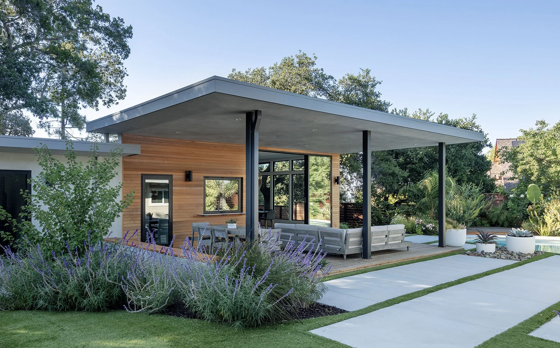 Modern house with outdoor patio, surrounded by greenery, featuring a flat roof, glass doors, and potted plants.