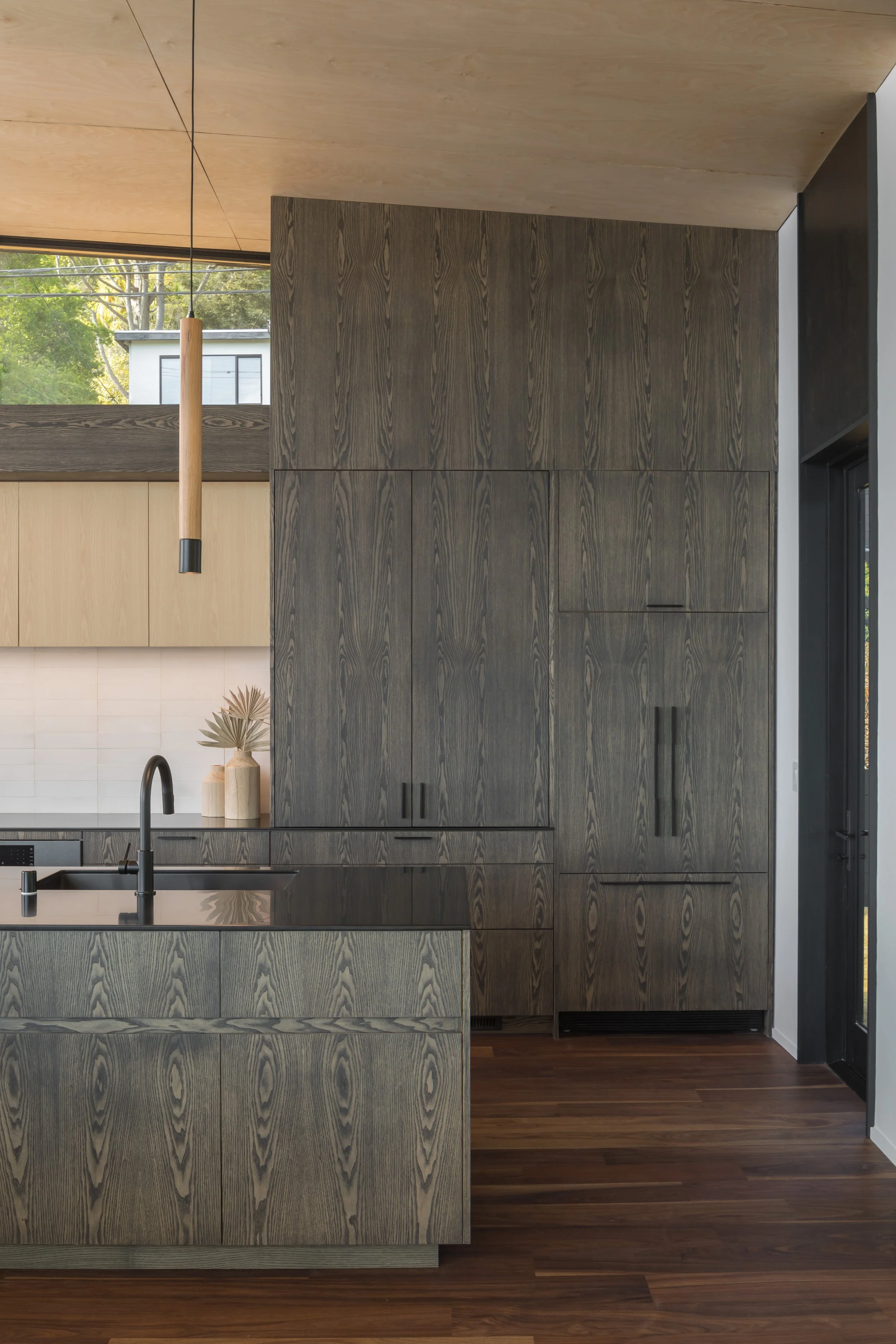 Interior view of a modern kitchen with dark wood cabinetry, black fixtures, and a wooden ceiling. Large window shows green trees outside, and there is a black door to the right. A pendant light hangs from the ceiling.