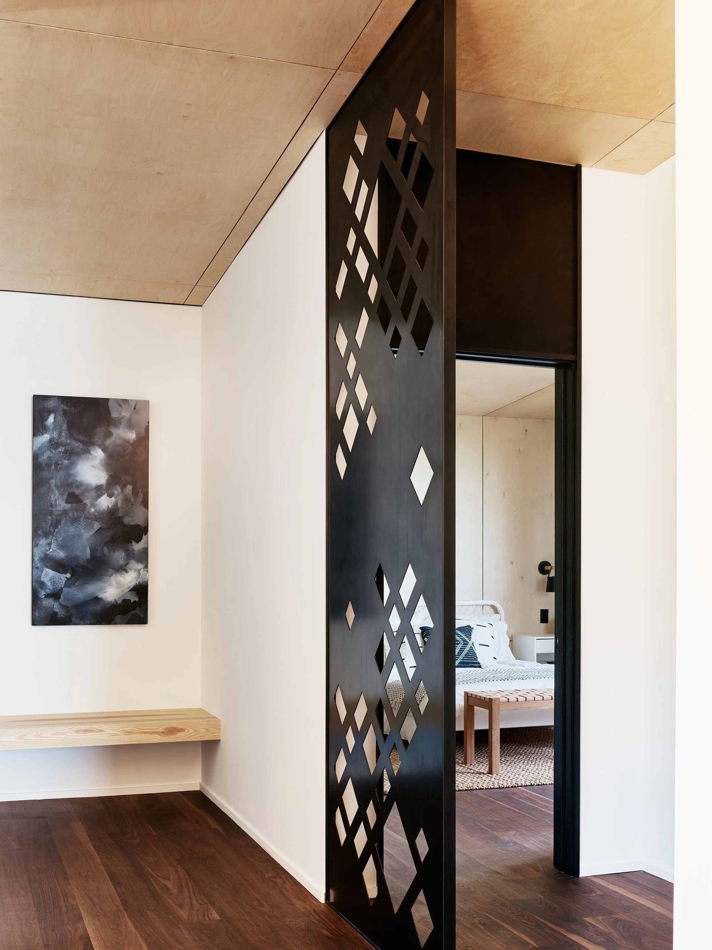 Interior view of a modern living space with a black decorative partition, a wooden bench, abstract art on a white wall, and a bedroom visible through an open doorway.
