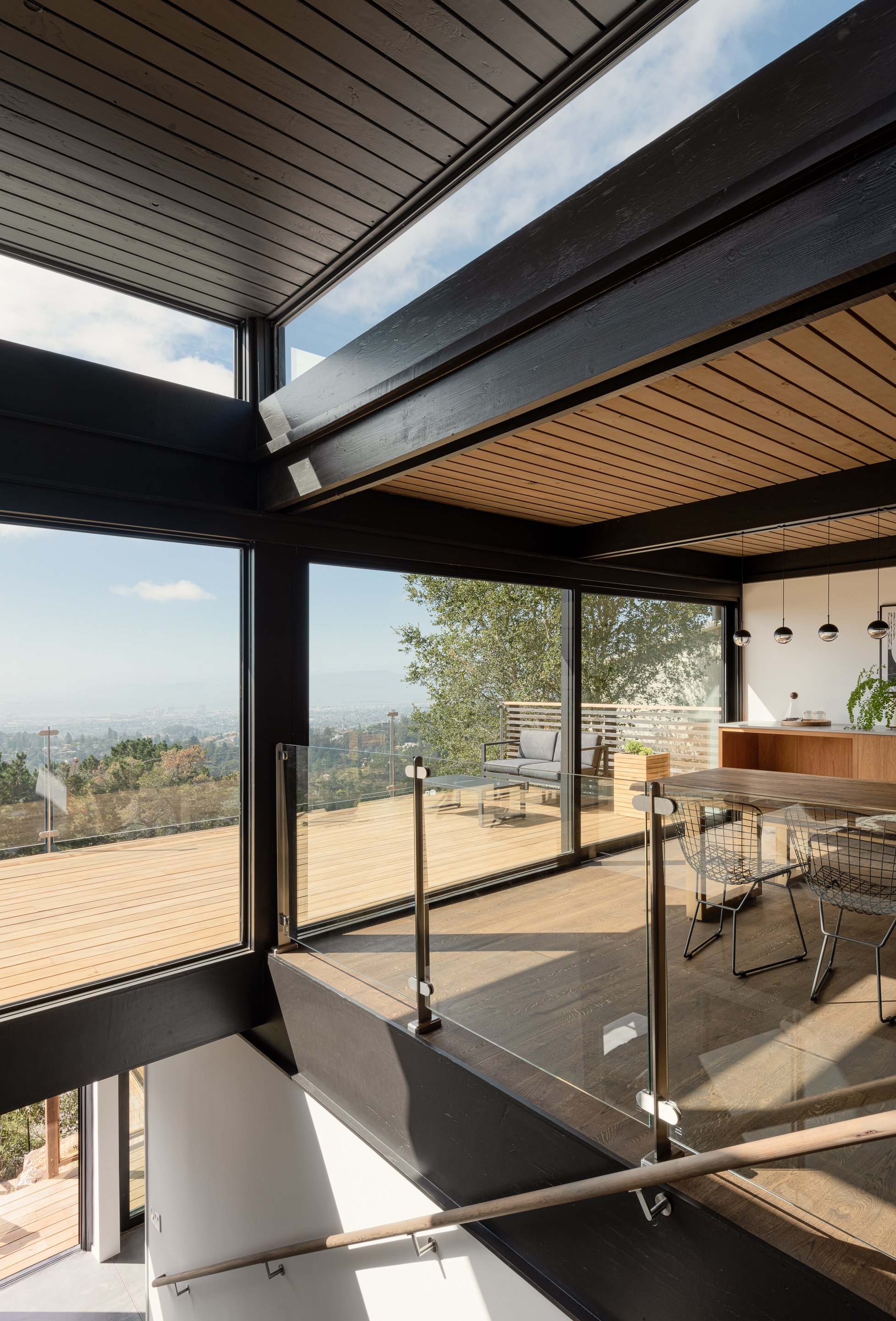 Interior view of a modern house with large glass windows and wooden ceiling and flooring. There is an outdoor deck with wooden flooring and patio furniture visible through the windows. The room has hanging pendant lights and a view of greenery and sk