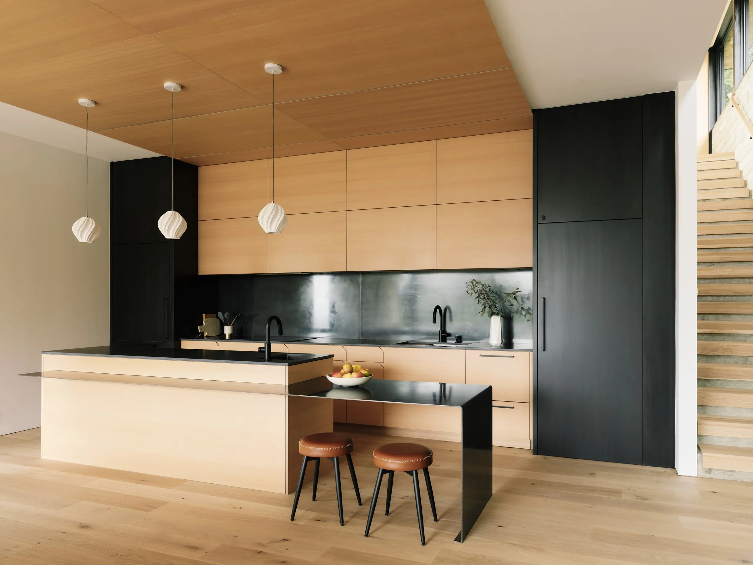 Modern kitchen with light wood cabinets and black accents, featuring a large kitchen island with a cream front and black countertop, hanging pendant lights, a bowl of apples, and a staircase with wooden steps leading upwards.