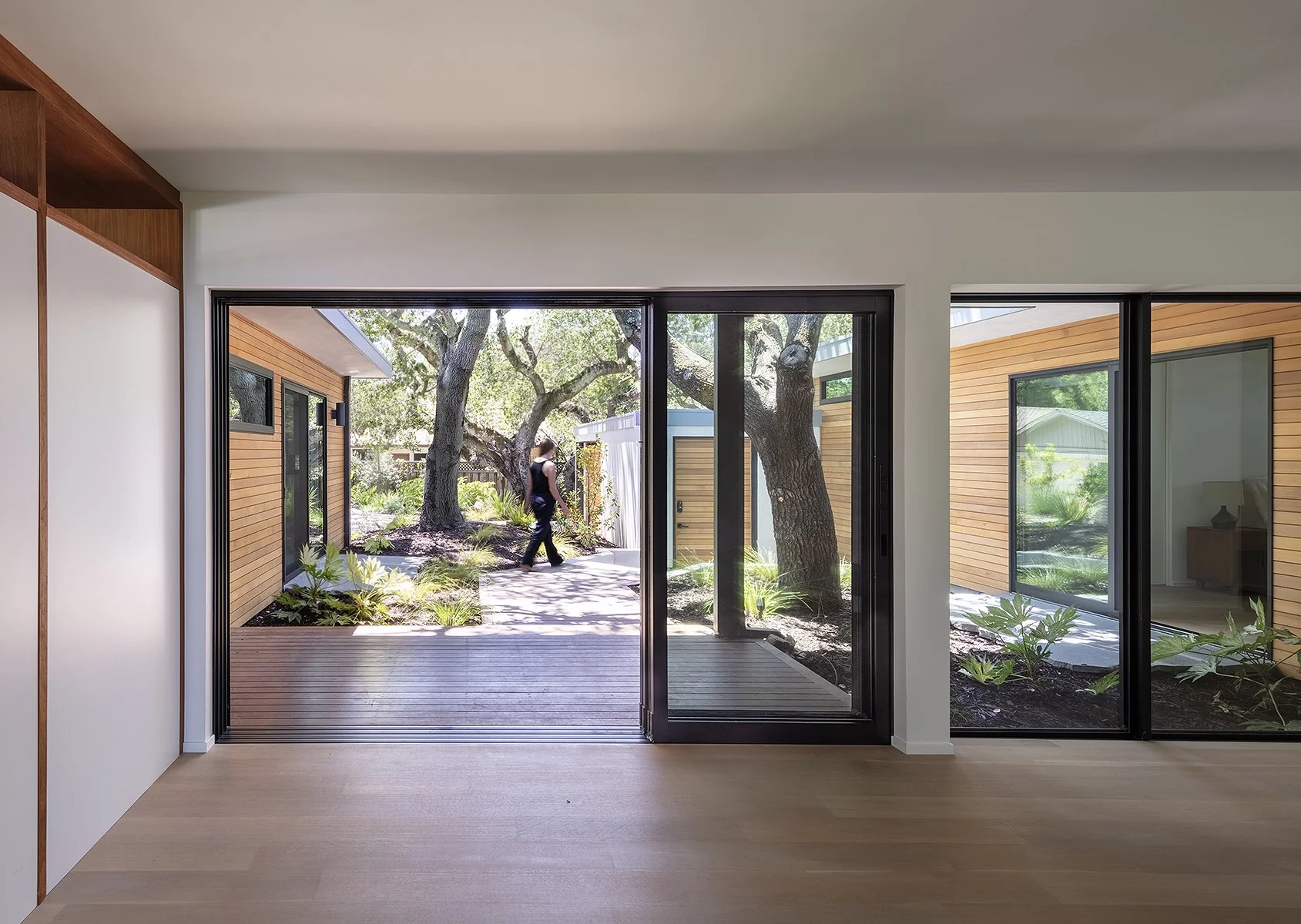 Interior view of a modern home with large sliding glass doors opening to a garden with trees and plants; a person is walking outside.