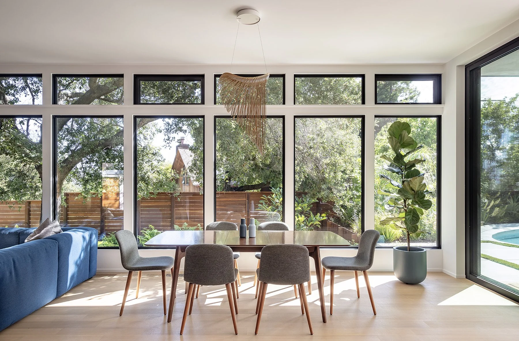 Interior of a modern living and dining area with floor-to-ceiling windows showcasing green trees and a backyard fence. There is a wooden dining table with seven gray chairs, a large green potted plant, and a blue sofa with pillows. A decorative penda