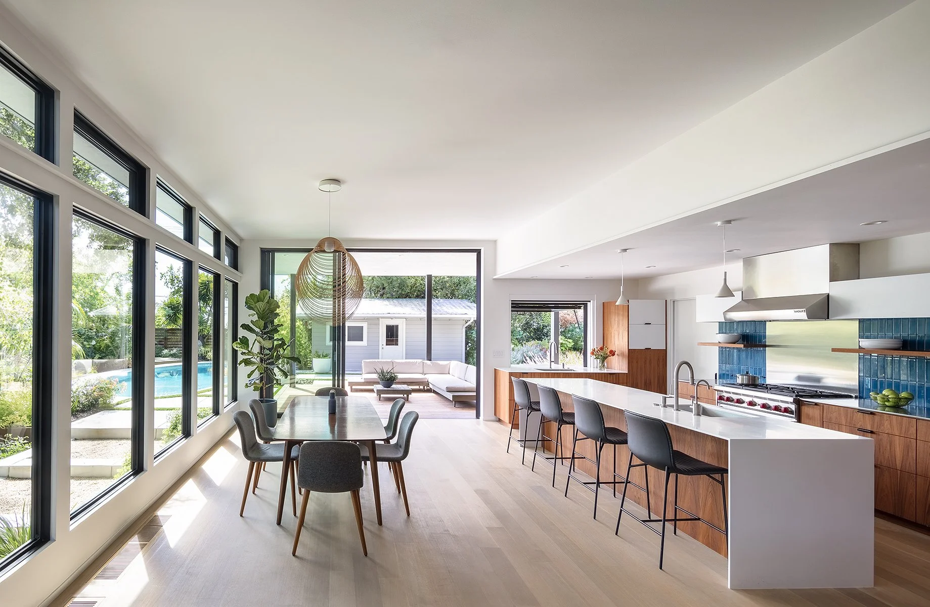 Open concept kitchen and dining area with large windows, patio doors, modern wood cabinetry, white countertops, blue tiled backsplash, kitchen island with black bar stools, and a living room with a white sofa and large potted plants outside.