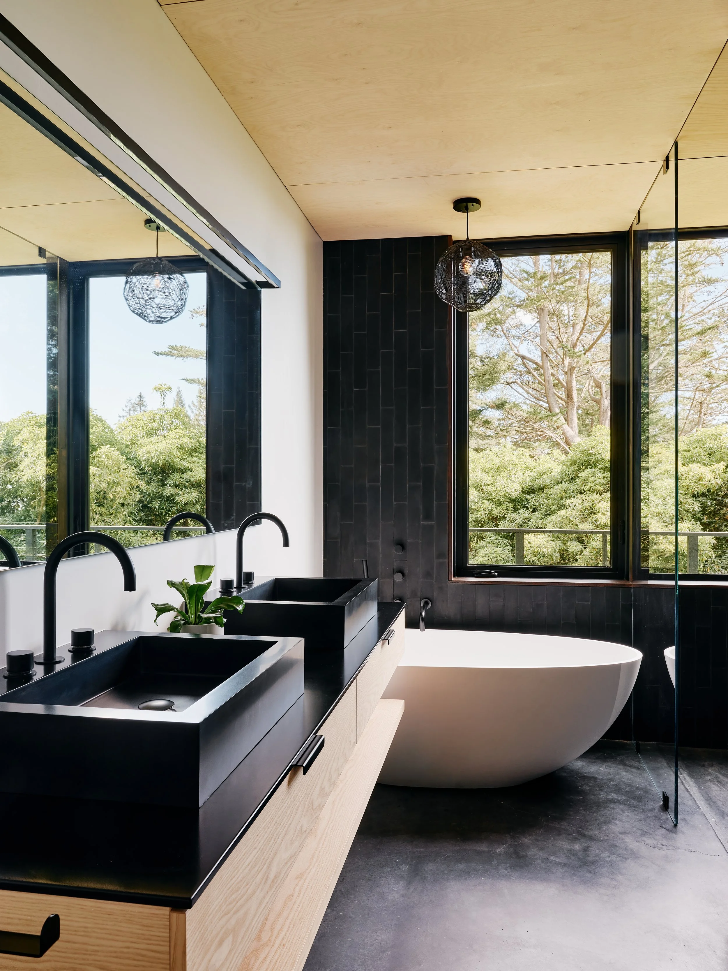 Modern bathroom with black tile and a large window showing trees outside. It includes black double sinks with wooden cabinetry, a white freestanding bathtub, and geometric pendant lights.