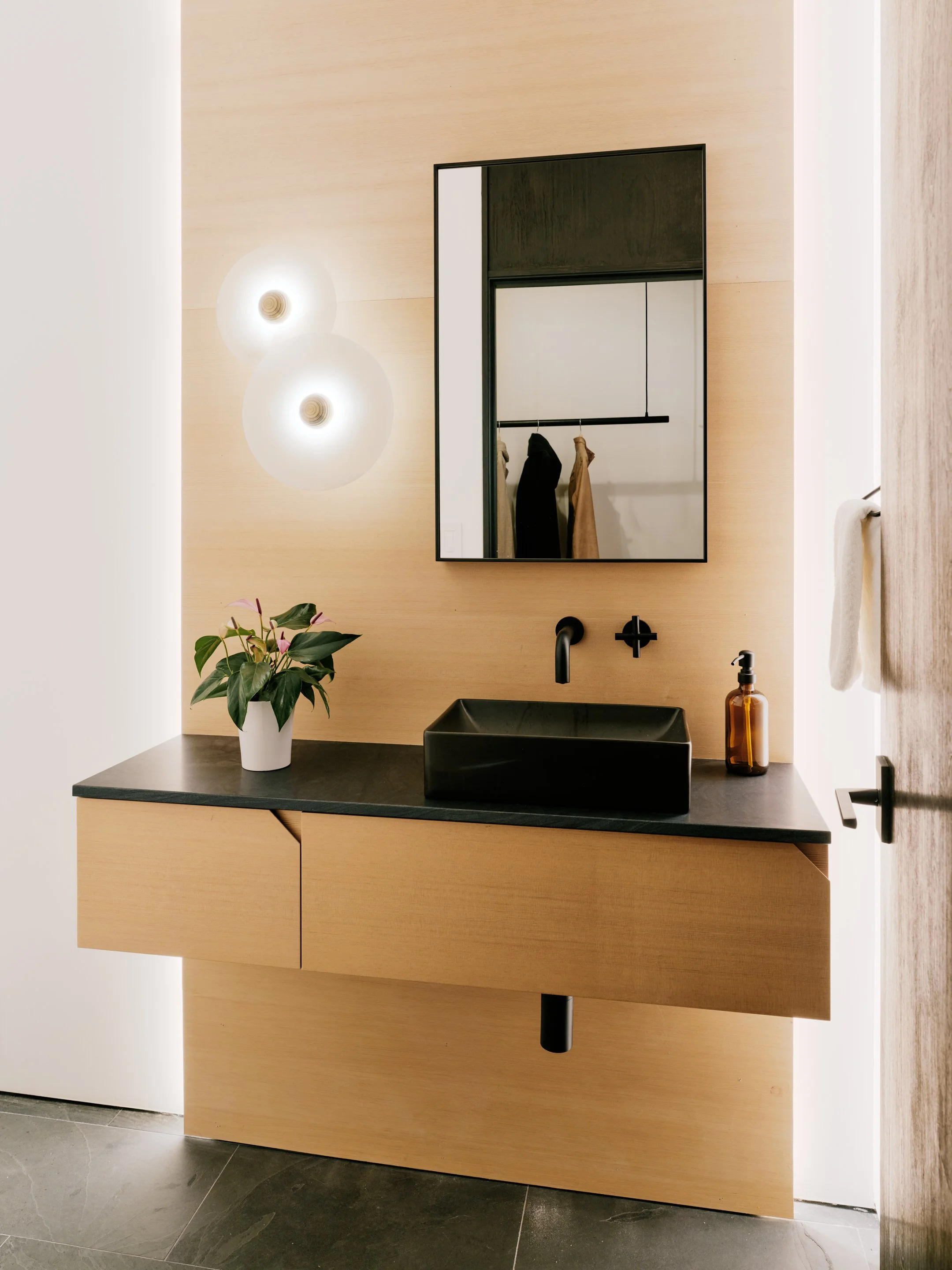 A modern bathroom vanity with a black vessel sink, a wall-mounted black faucet, a brown glass soap dispenser, a white planter with a green plant, and a rectangular mirror on a light wood wall.