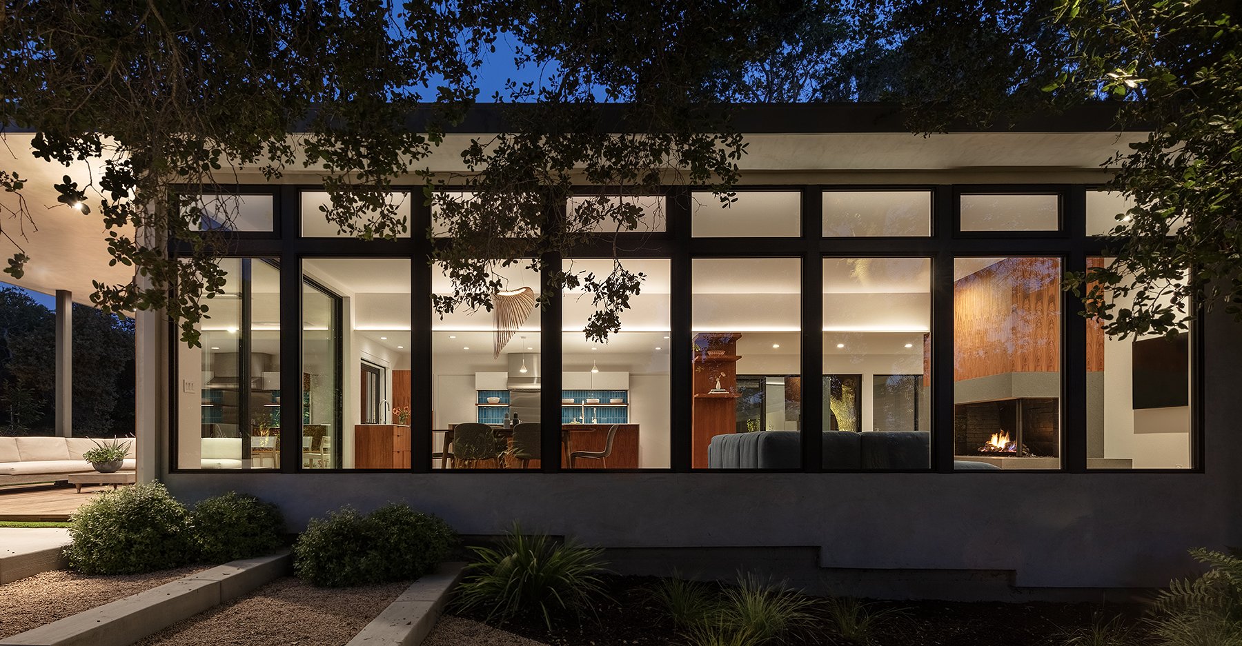 Modern house with large glass windows at dusk, inside living room and kitchen visible, fireplace burning, outdoor patio with seating and landscaping.