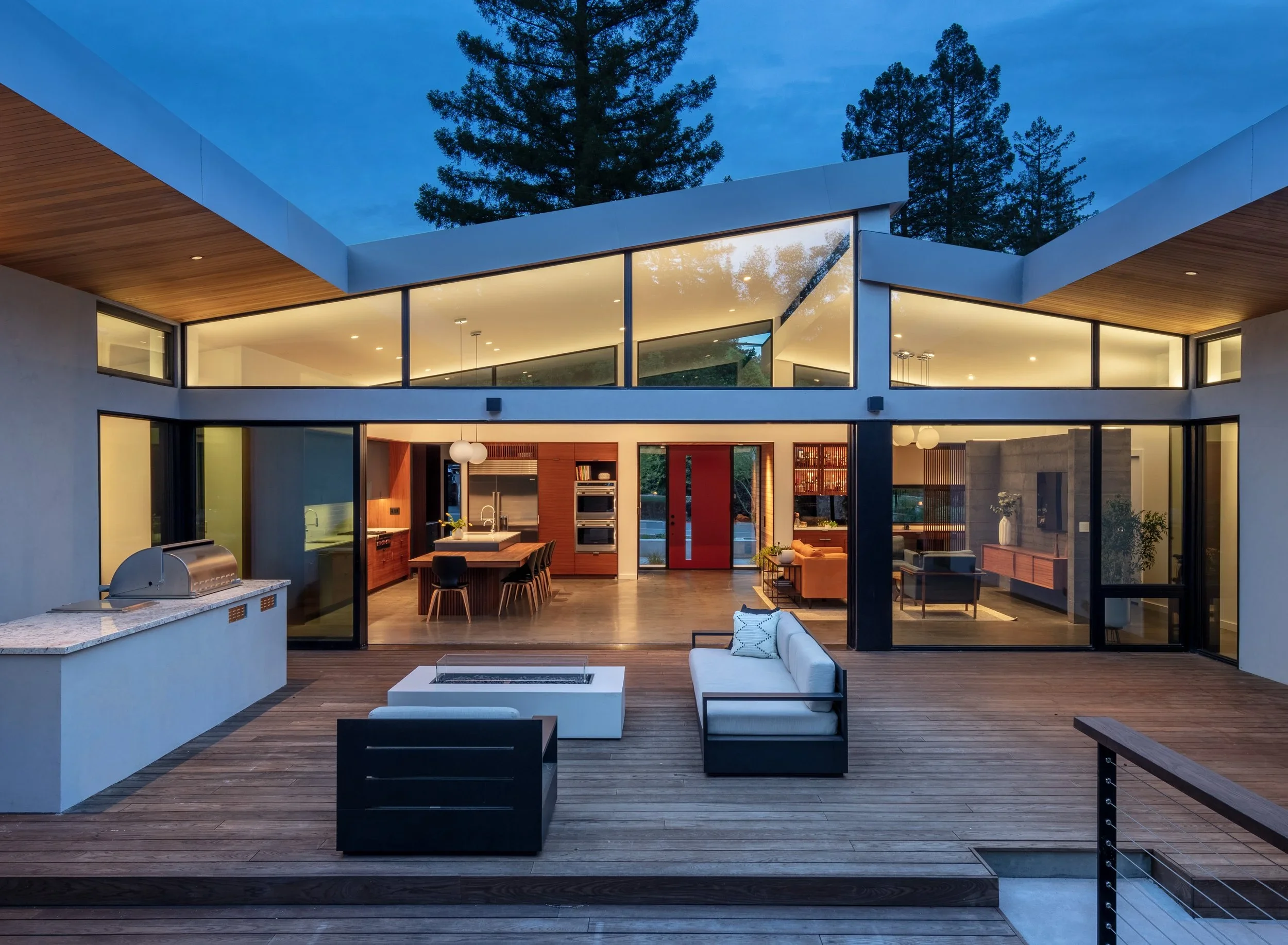 Modern two-story house with large glass windows and an outdoor wooden deck, illuminated at dusk, surrounded by tall pine trees.