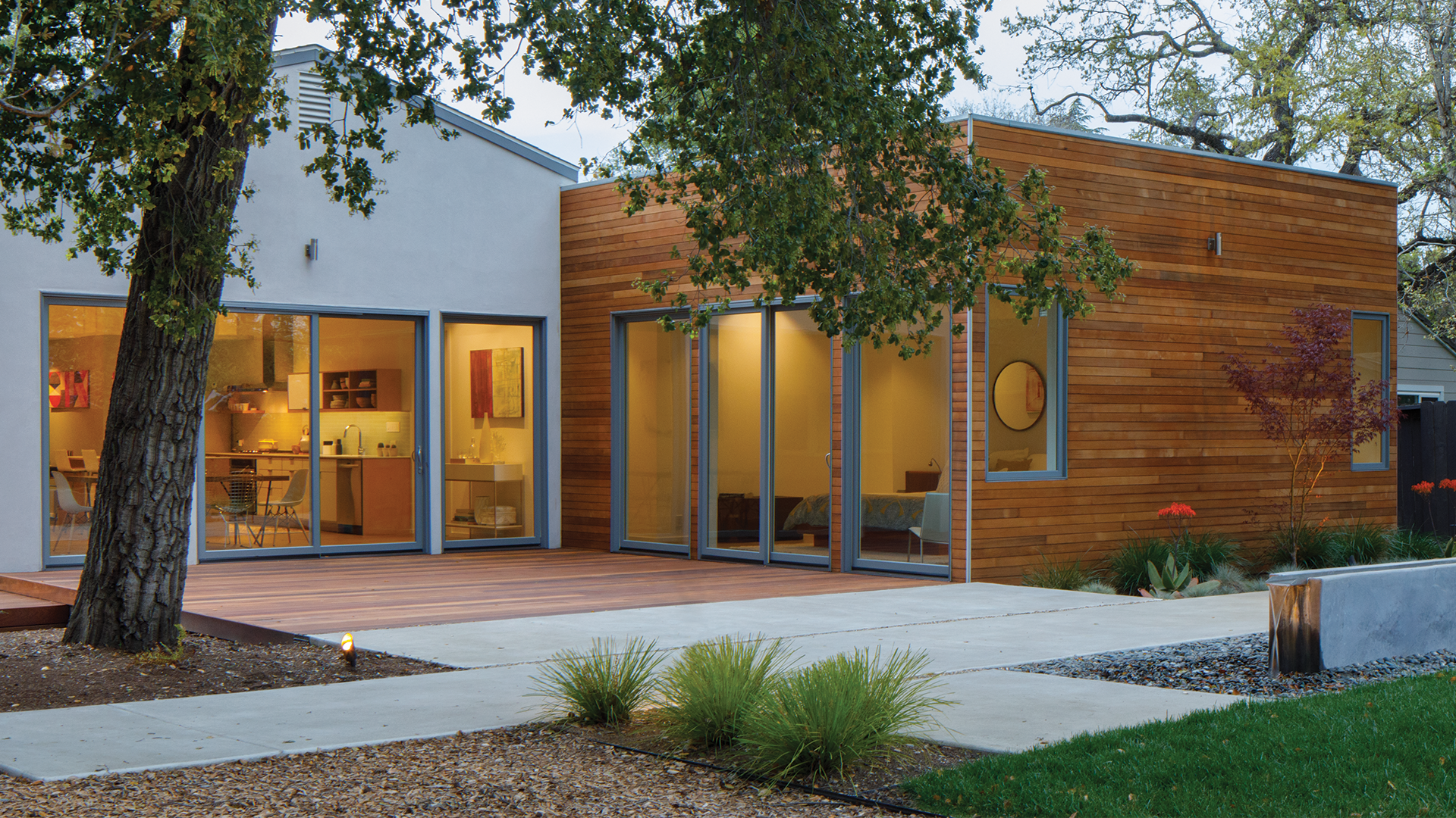Modern house with wooden and white exterior panels, large glass sliding doors, and a backyard with concrete pathway, landscaping, and trees