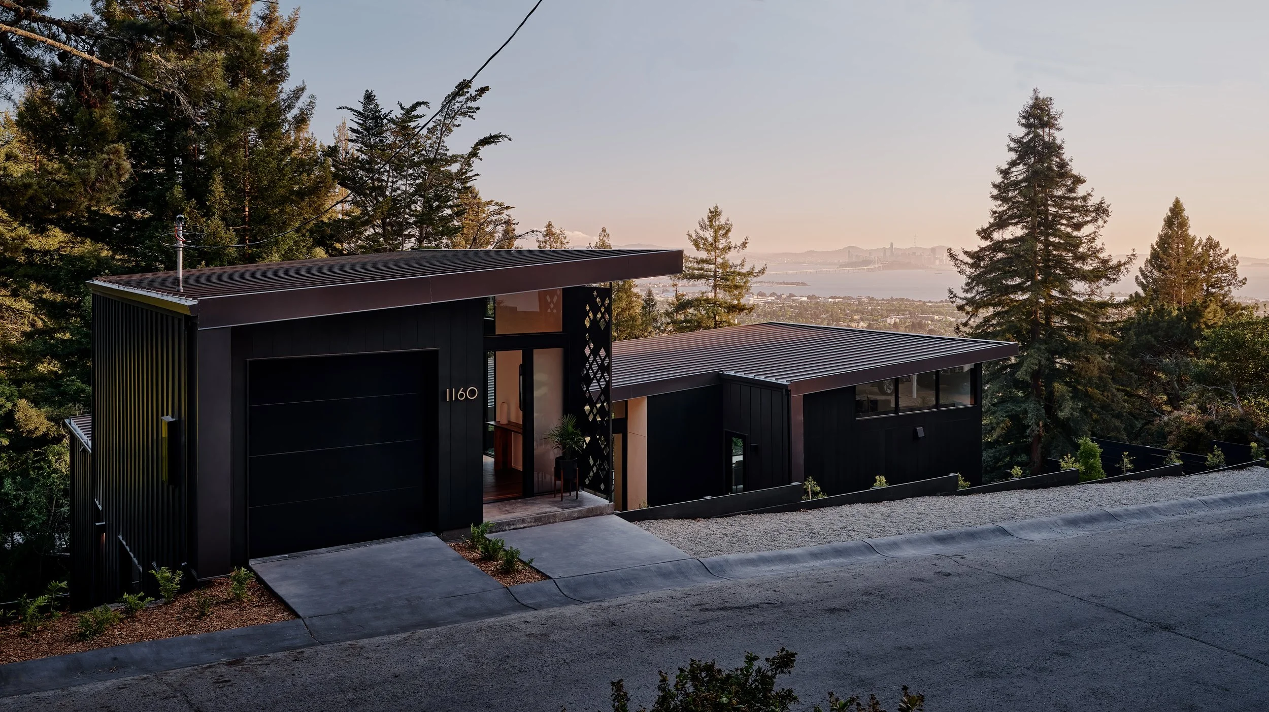 Modern black house on a hill surrounded by trees with a view of a body of water and distant city skyline in sunset