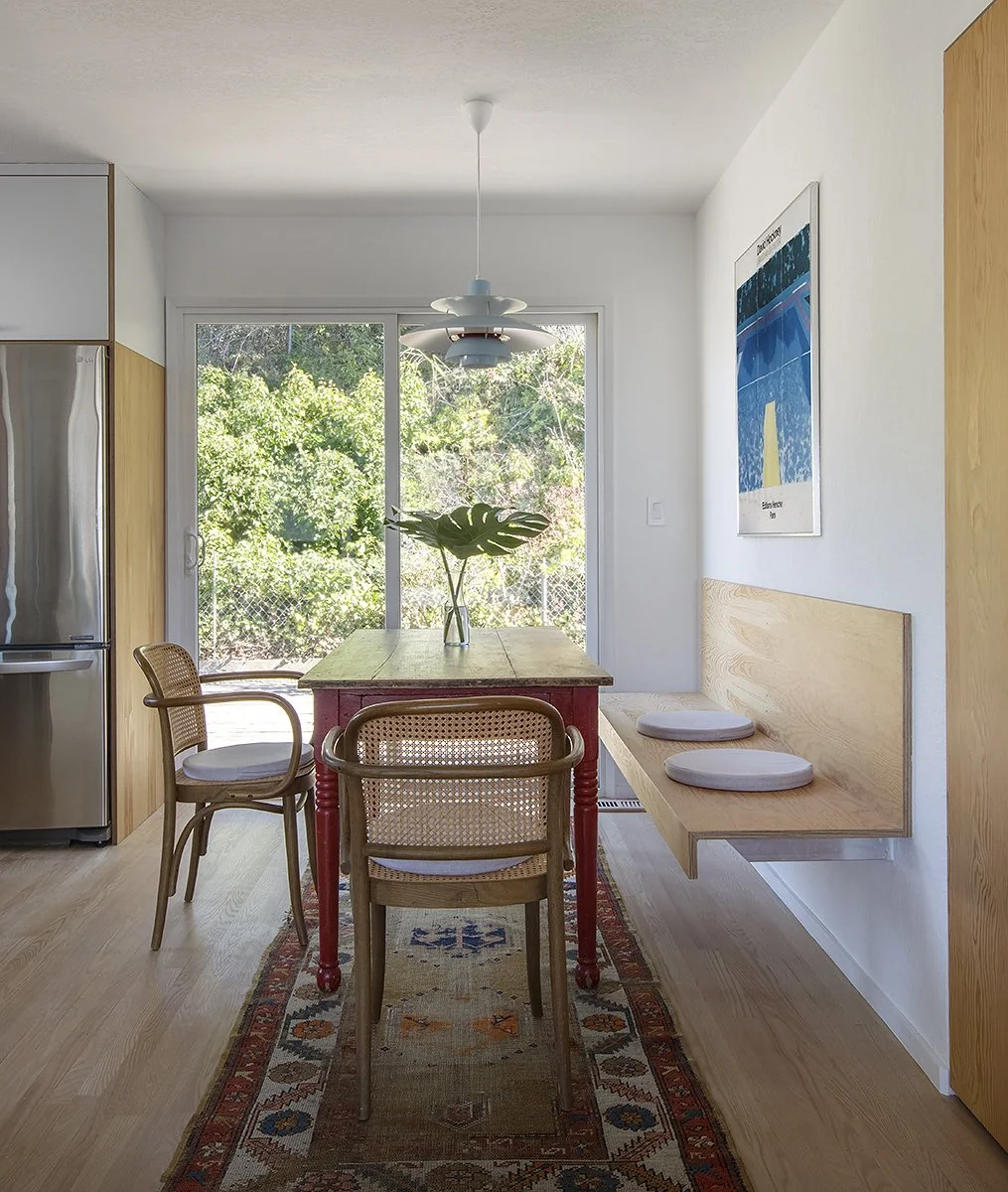 A dining room with a wooden table, three chairs, a bench with cushions, a hanging light fixture, a sliding glass door with greenery outside, and a framed poster on the wall.