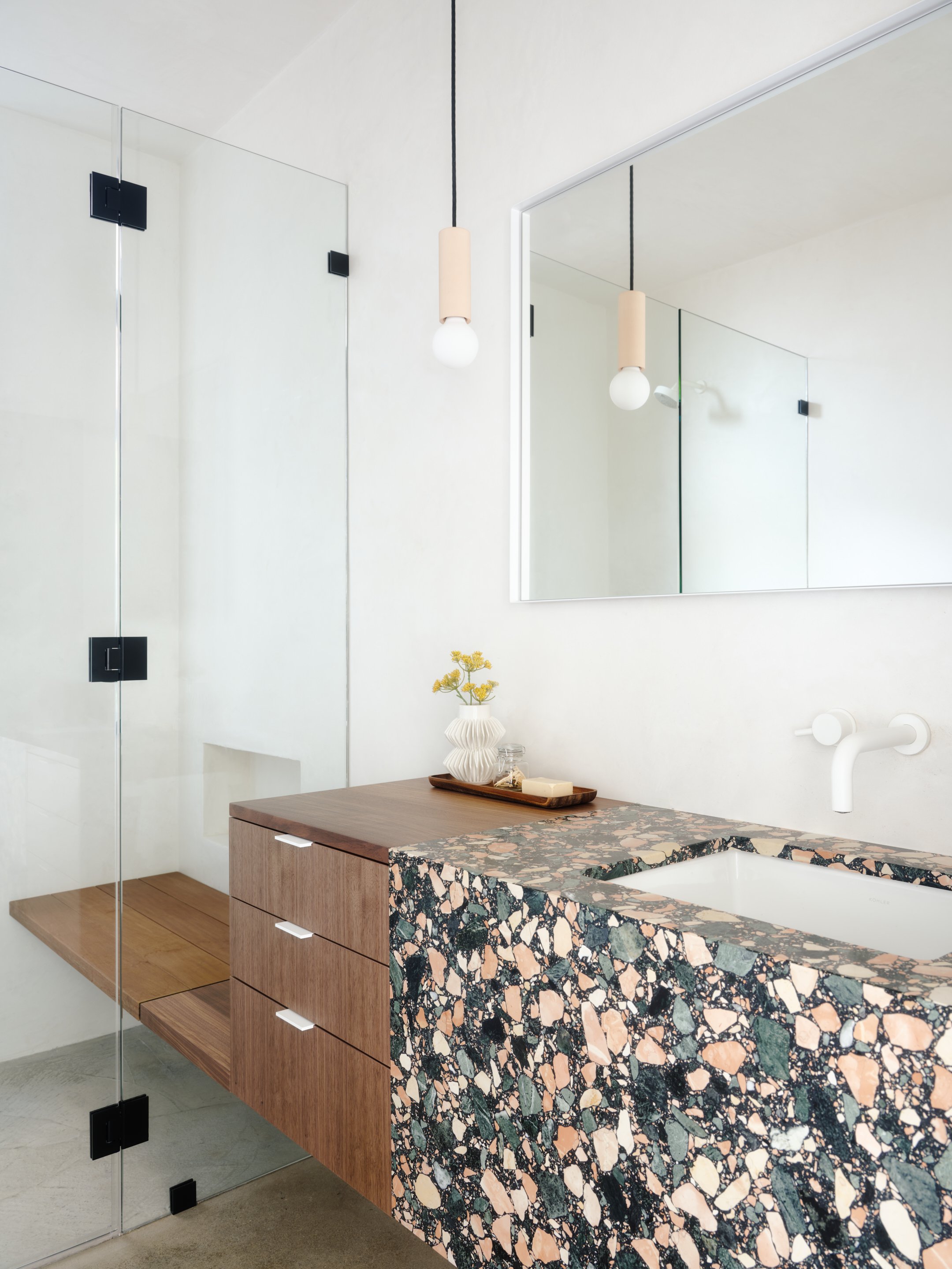 A modern bathroom vanity with a terrazzo countertop, a white sink, and wooden drawers, next to a glass shower enclosure with black hardware. There is a large mirror and a small vase with yellow flowers on the countertop.